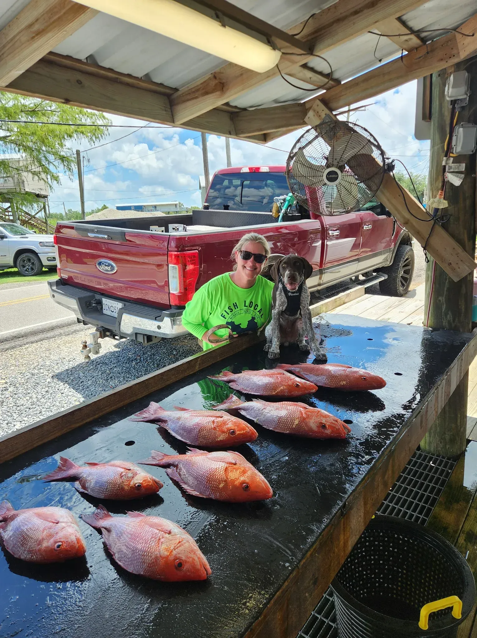 A person and a dog stand behind a cleaning table covered in several red fish, with a red truck parked in the background.
