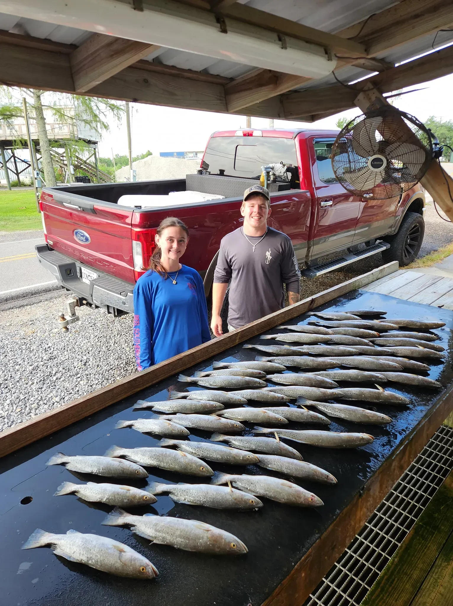 A person and a man stand behind a long cleaning table covered with dozens of freshly caught fish near a red pickup truck.