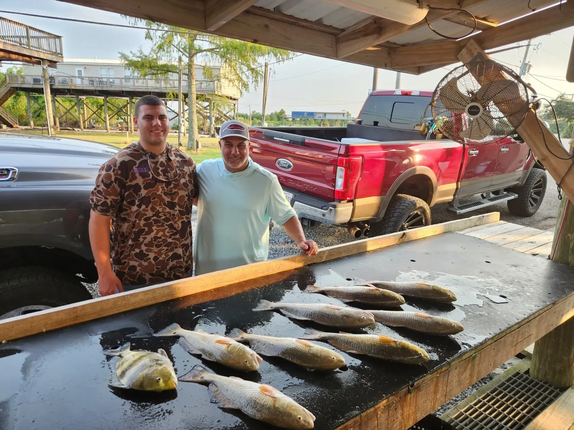 Two people smile behind a fish-cleaning table filled with freshly caught fish, with a red pickup truck parked nearby.