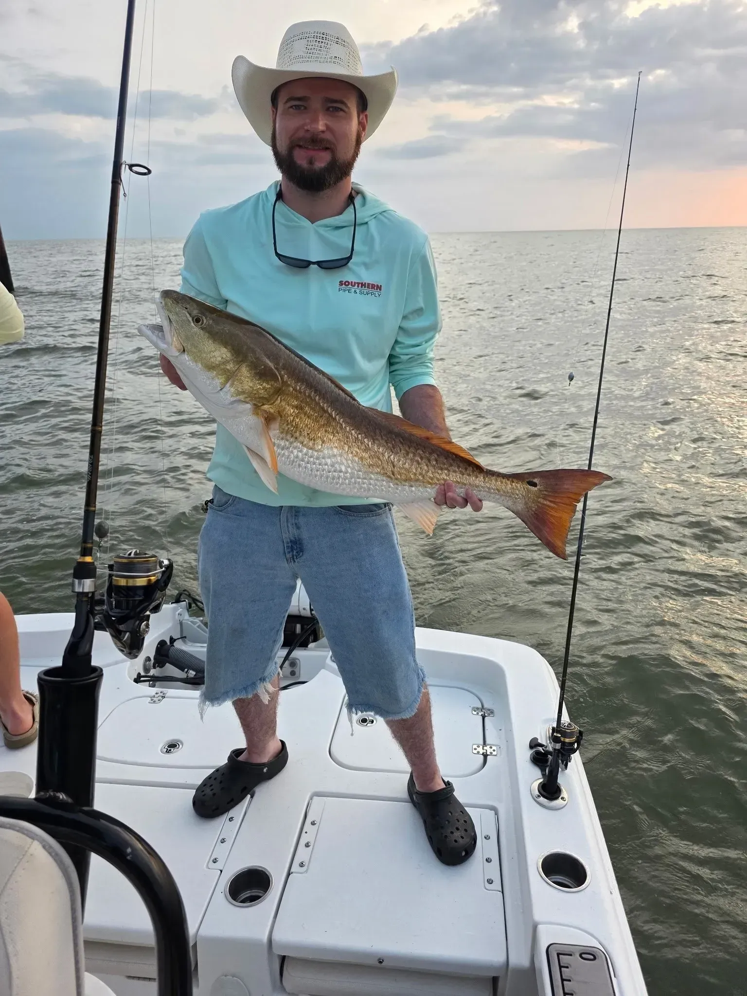 A person in a light blue long-sleeved shirt and cowboy hat holds a large red drum fish while standing on a boat.