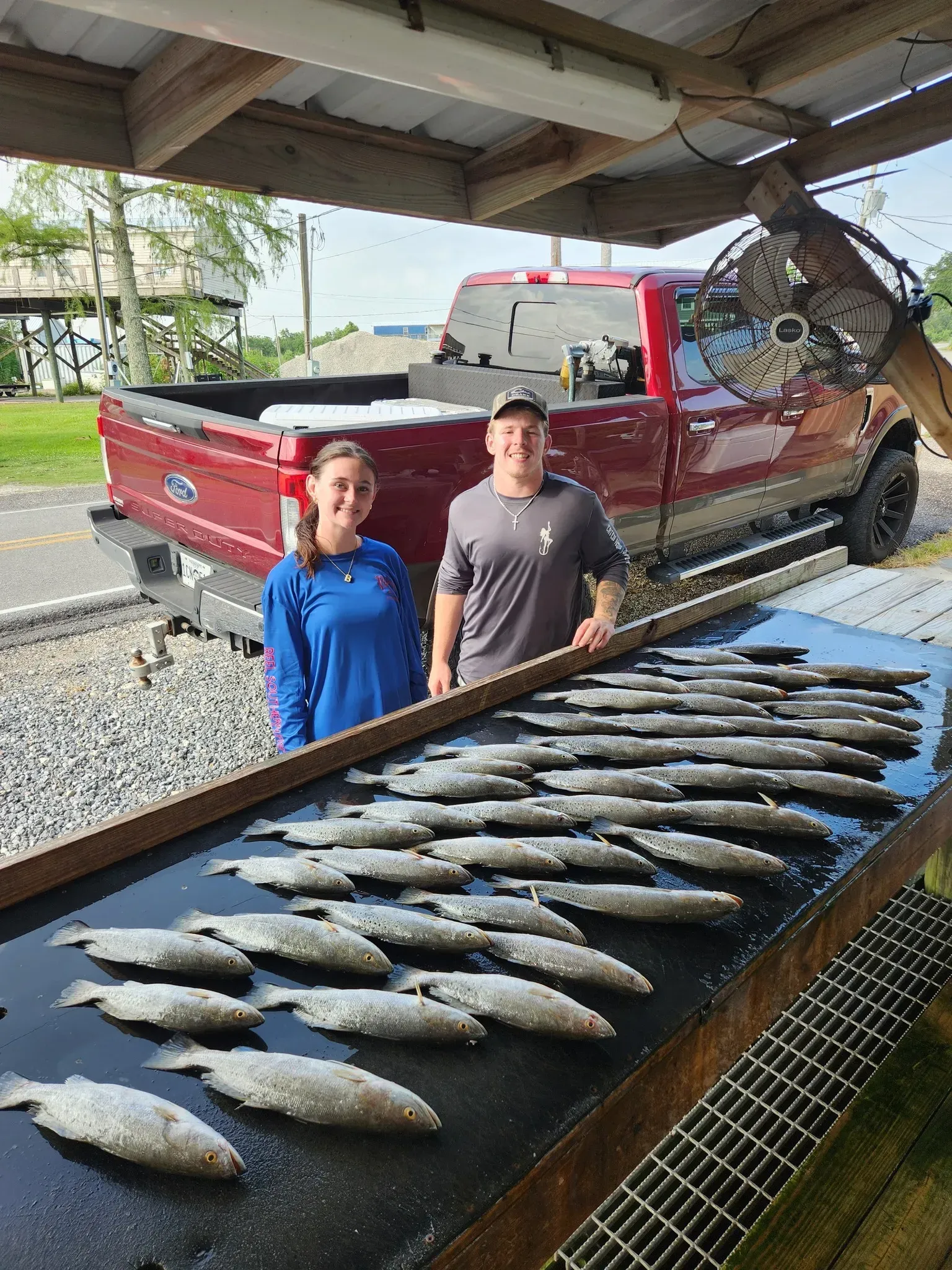 Two people stand behind a table filled with many freshly caught fish, with a red pickup truck parked in the background.