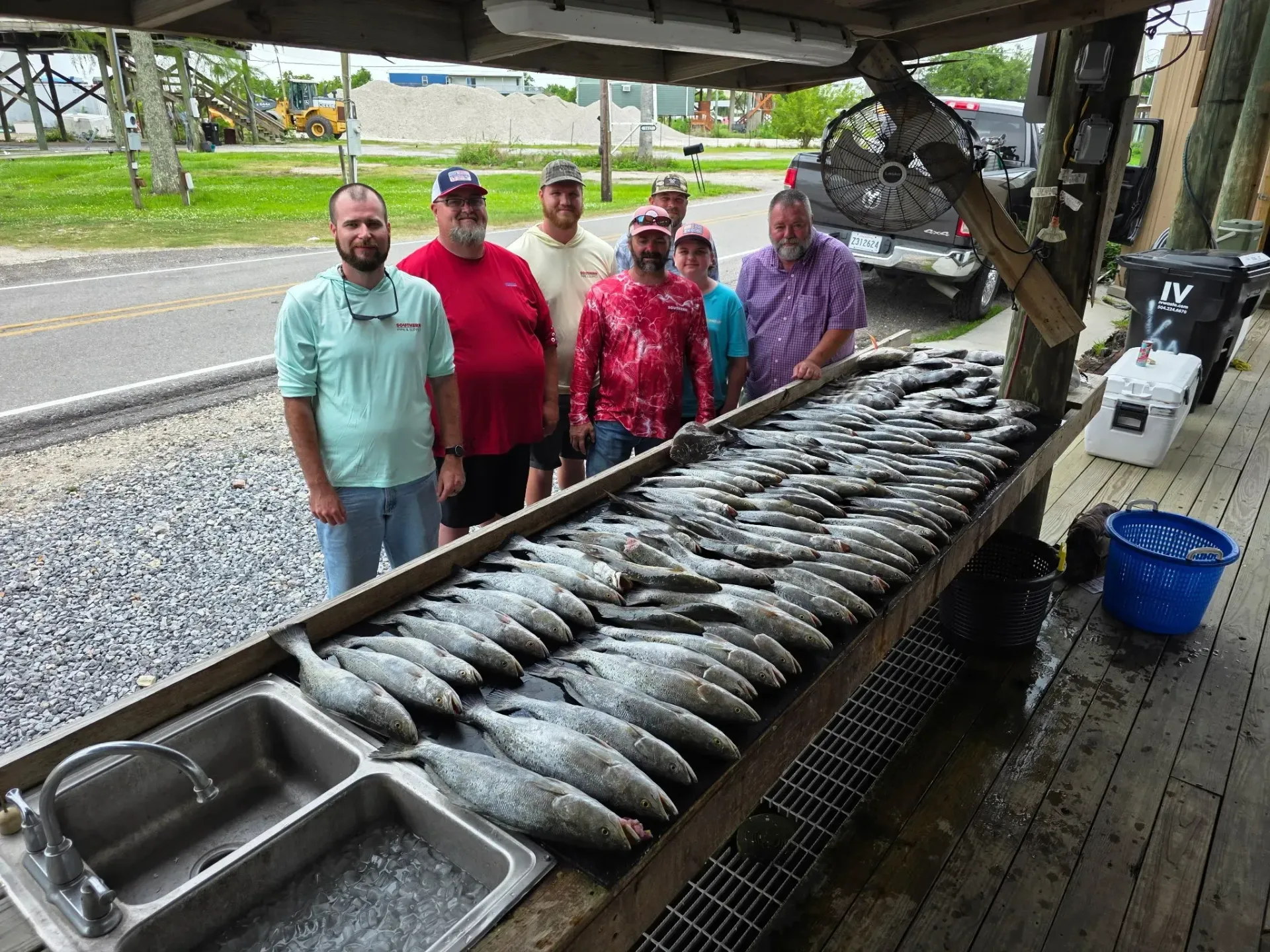 Seven people pose behind a long cleaning table filled with a large catch of freshly caught fish at an outdoor station.