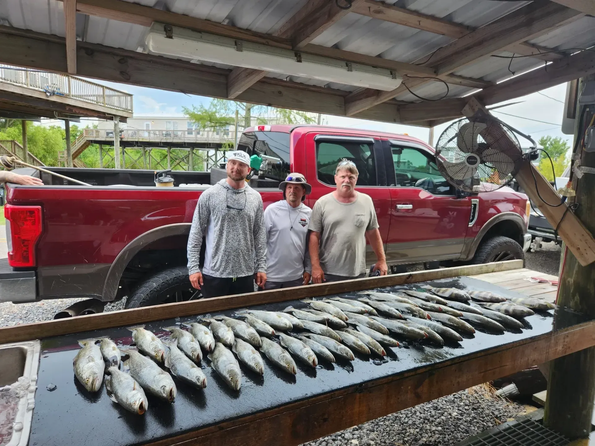 Three people stand behind a table covered in a large catch of fish, with a red truck parked in the background.