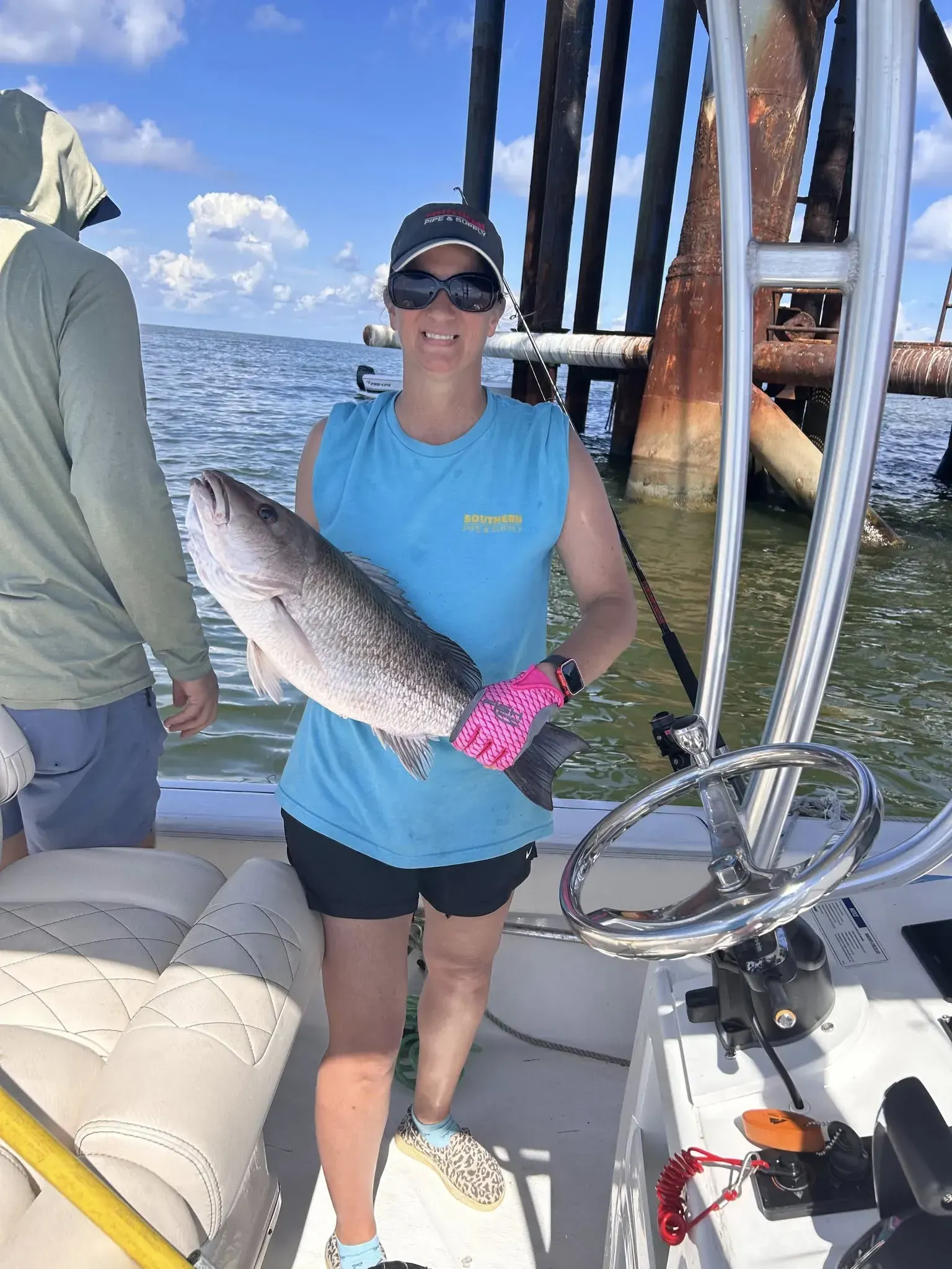 A person standing on a boat holds a large fish, wearing a blue shirt, sunglasses, a hat, and pink gloves.