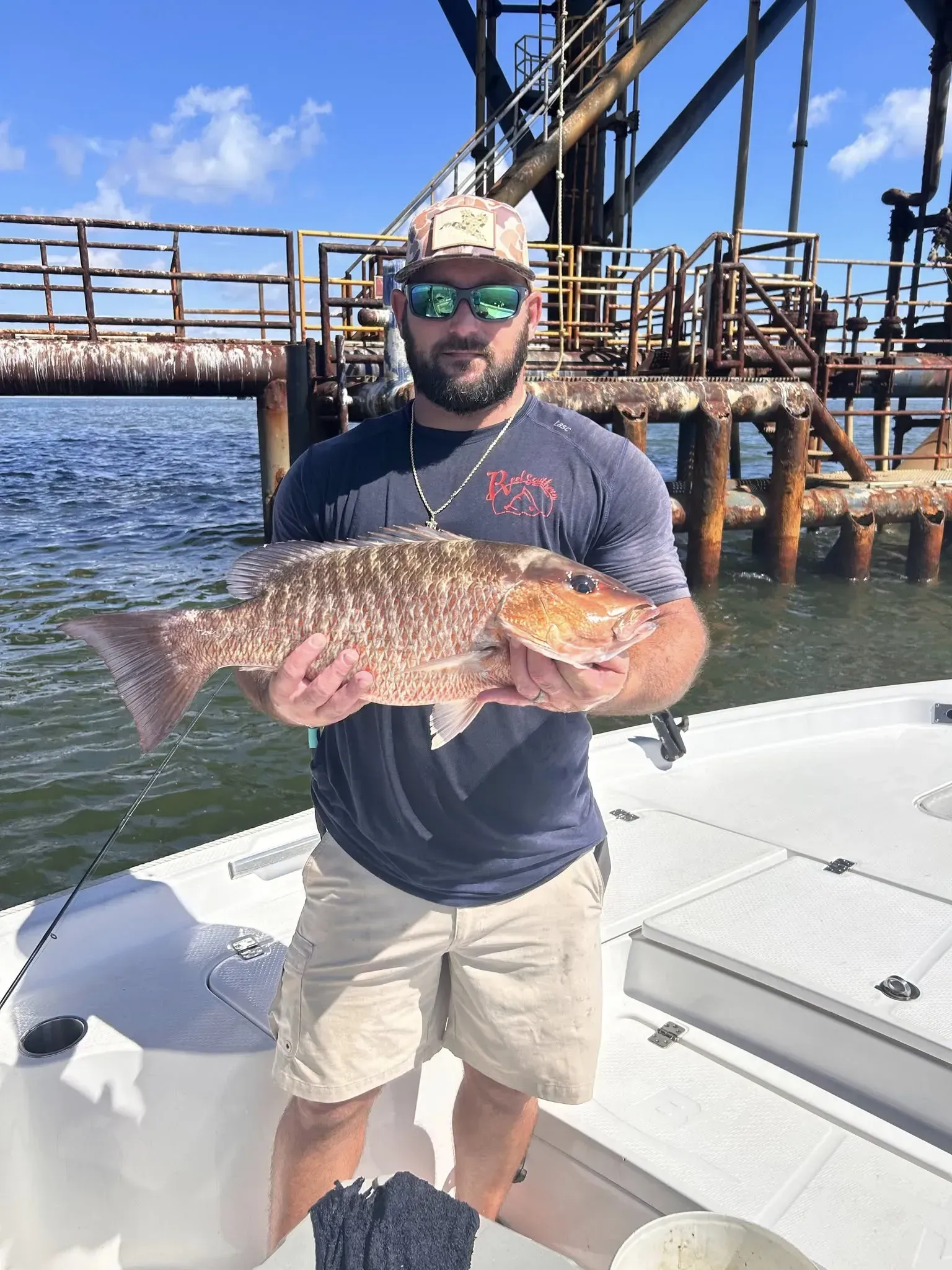 A man in a cap and sunglasses holds a large fish while standing on a boat in front of an offshore oil platform.