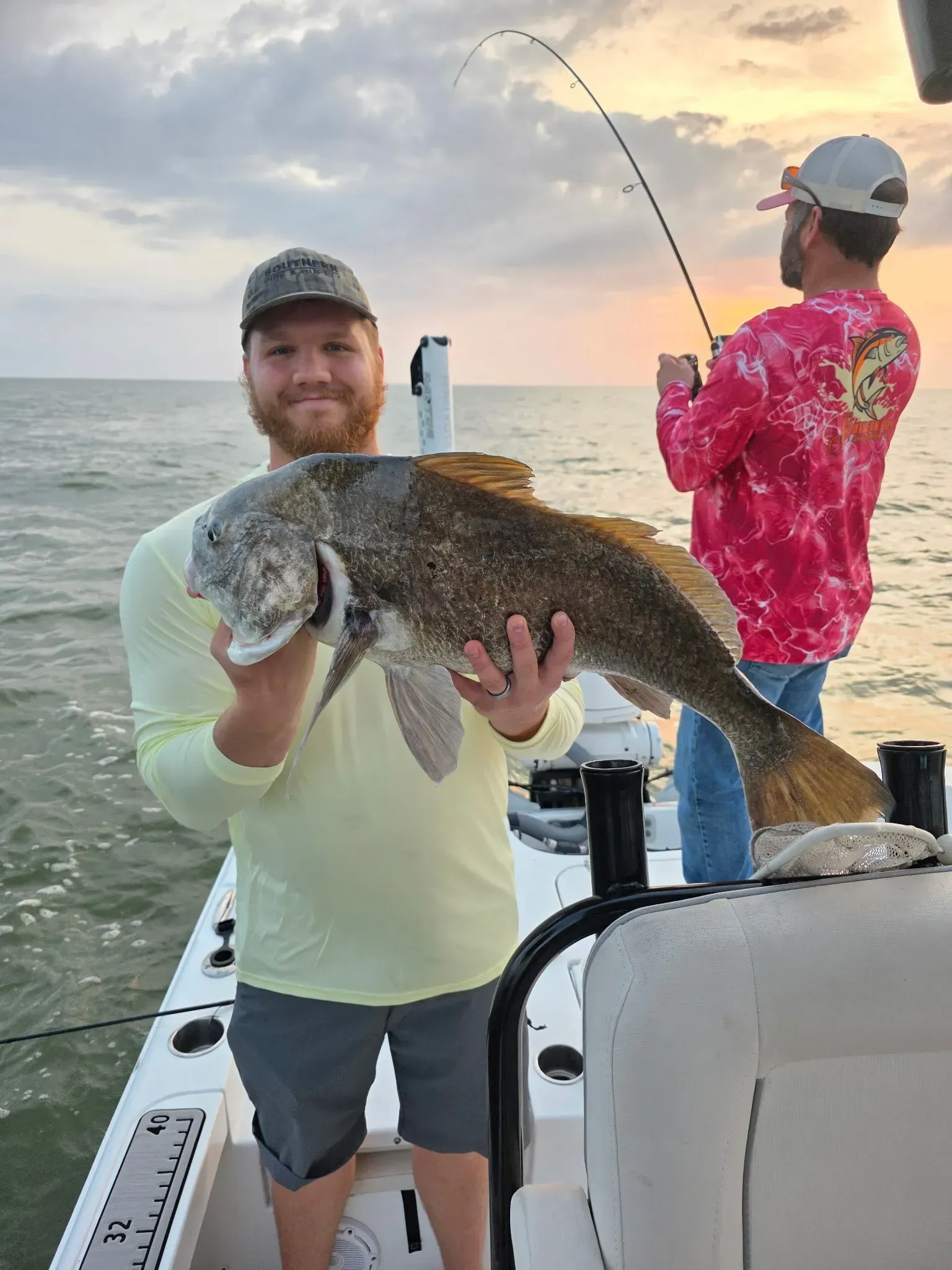A person holds a large fish on a boat while another person fishes in the background at sunset.
