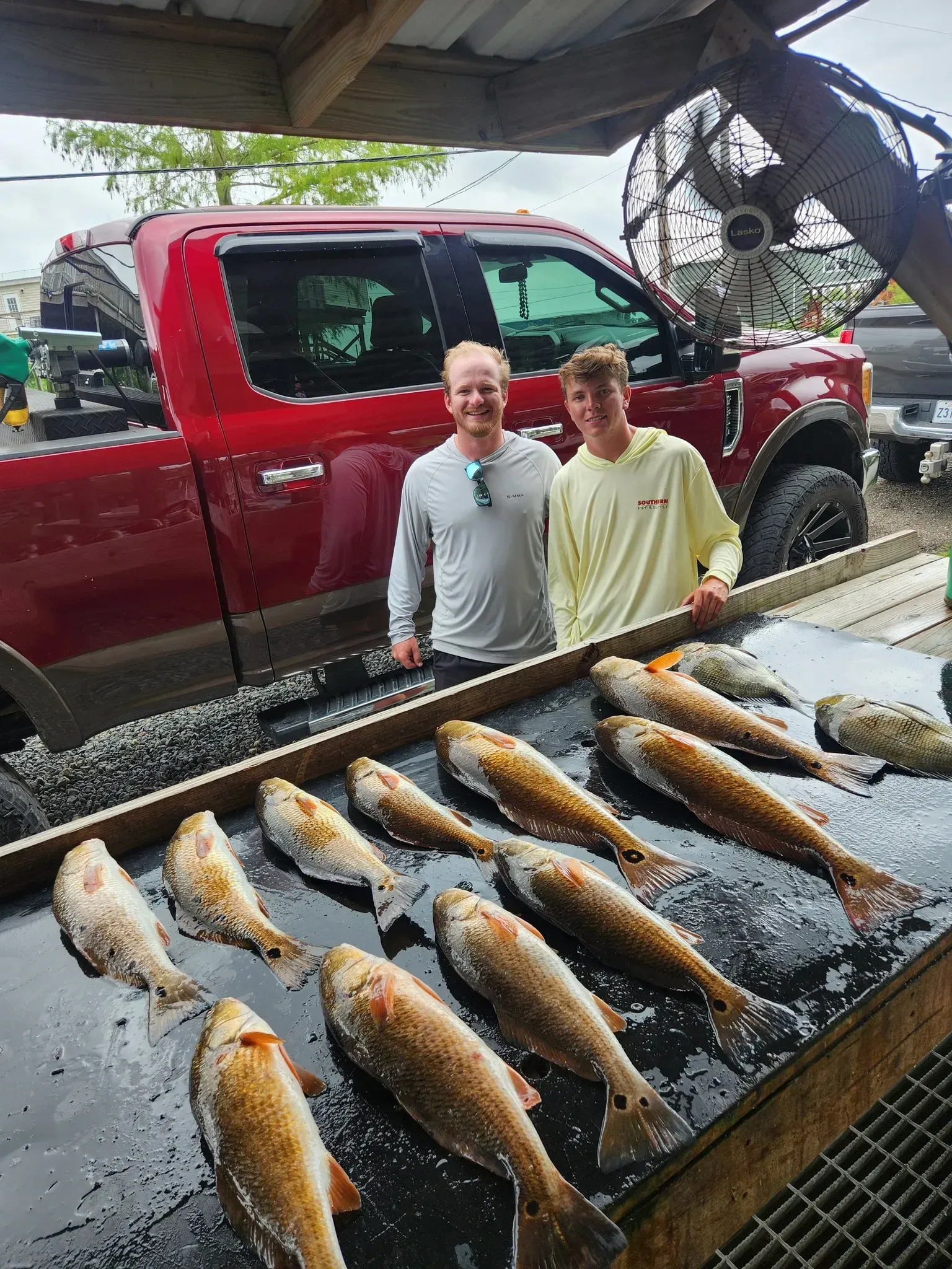 Two smiling people stand behind a fish cleaning station with several caught fish, in front of a bright red pickup truck.