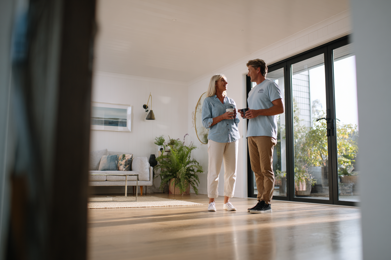 Two people talking in a sunlit room. Woman in blue shirt and man in a blue polo holding mugs, smiling. Large windows.