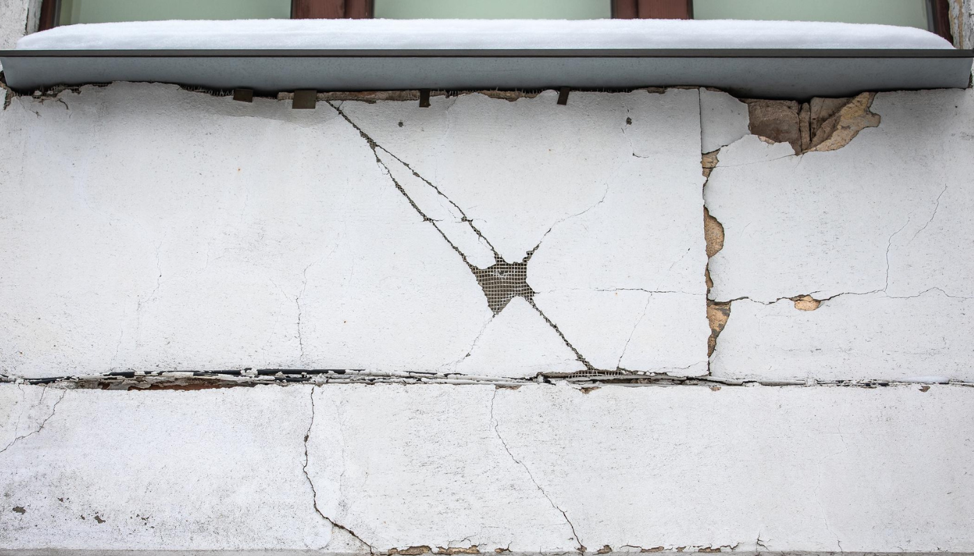 Cracked, deteriorating white stucco wall beneath a window, with visible damage and crumbling sections.