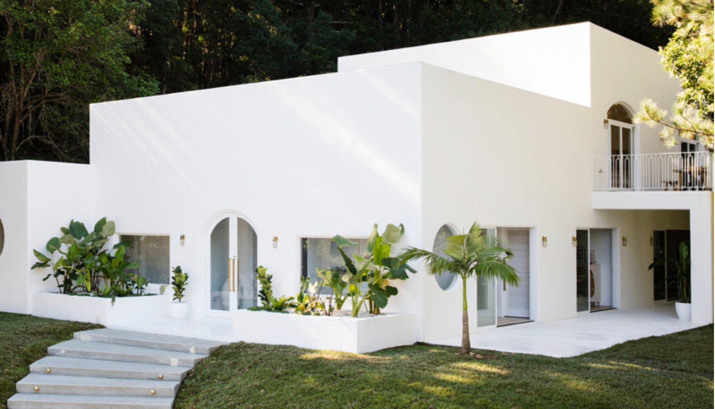 White, modern house with arch details and landscaping, set amidst green trees and grass.
