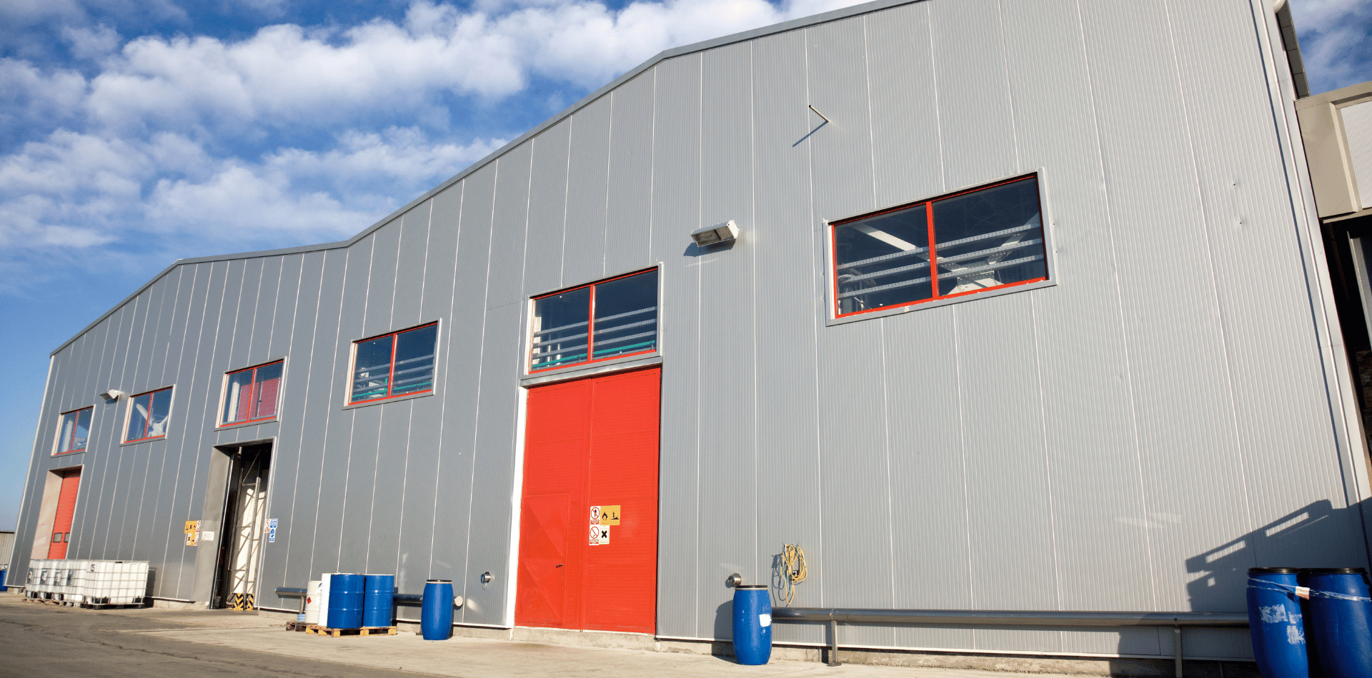 A warehouse exterior with light gray corrugated metal siding, several rectangular windows, and a large red loading door.
