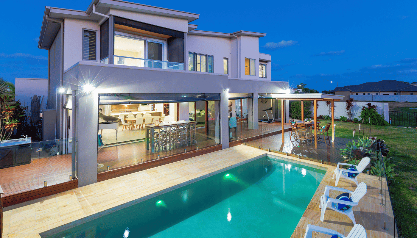 A two-story modern house at twilight with a lit swimming pool, a patio, and three white chairs on a paved deck.