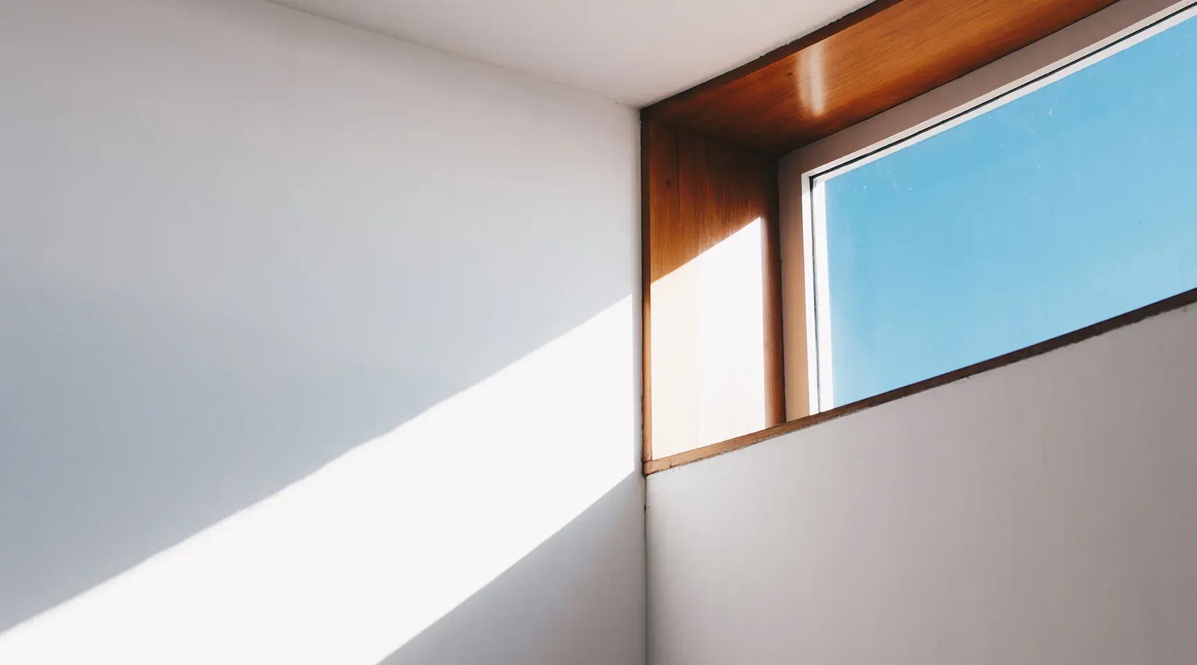 White room corner with wooden-framed window, blue sky visible, bright sunlight casting shadows.