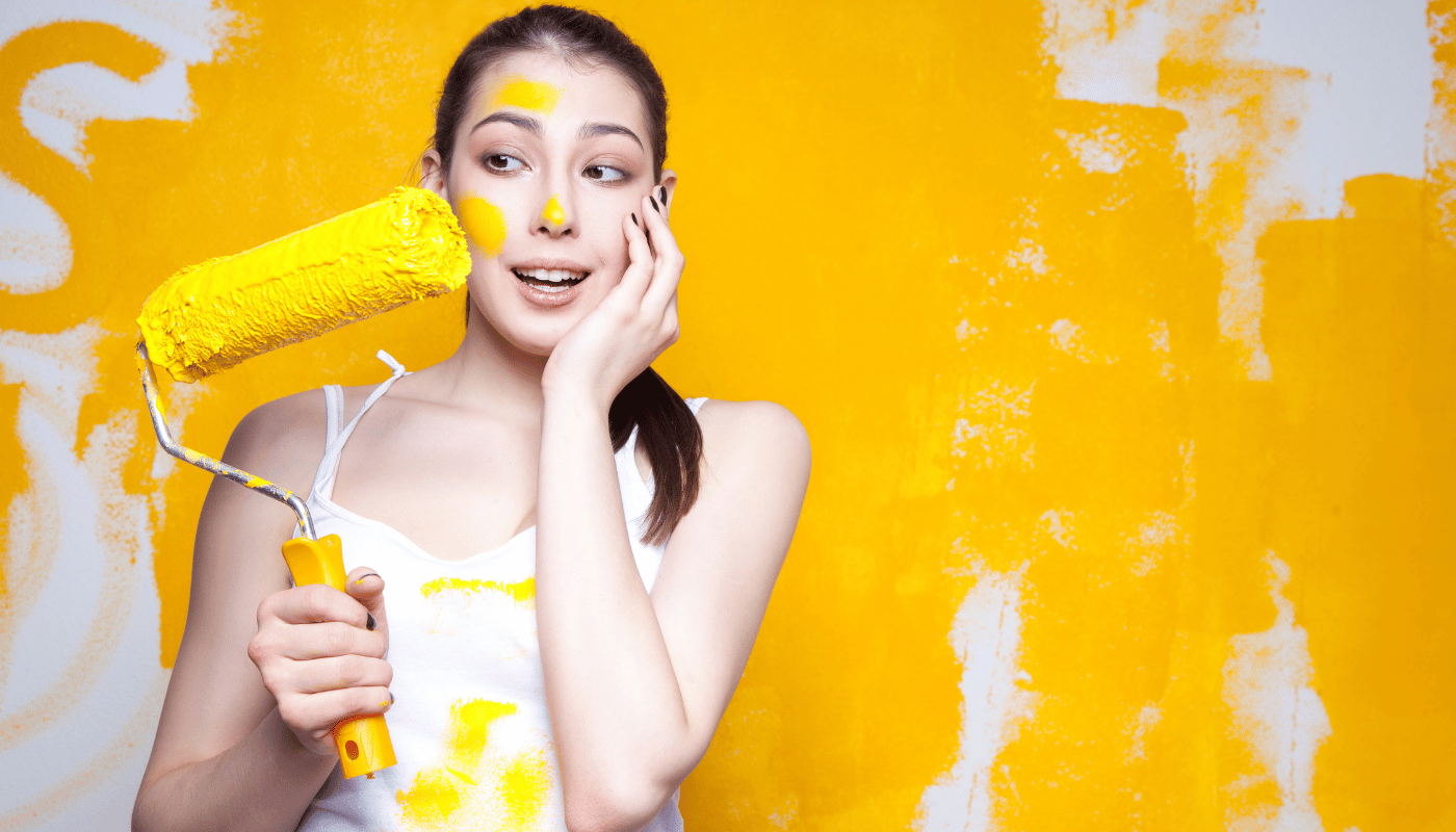 A woman is holding a paint roller with yellow paint on her face.