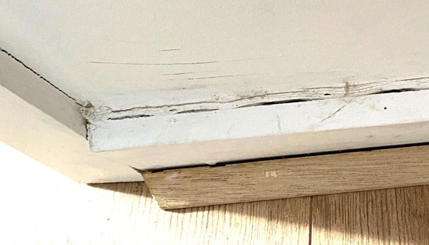 Close-up of a cracked, white ceiling with dark lines, revealing underlying wood trim.