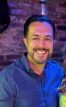 A man with a beard is smiling in front of a brick wall.