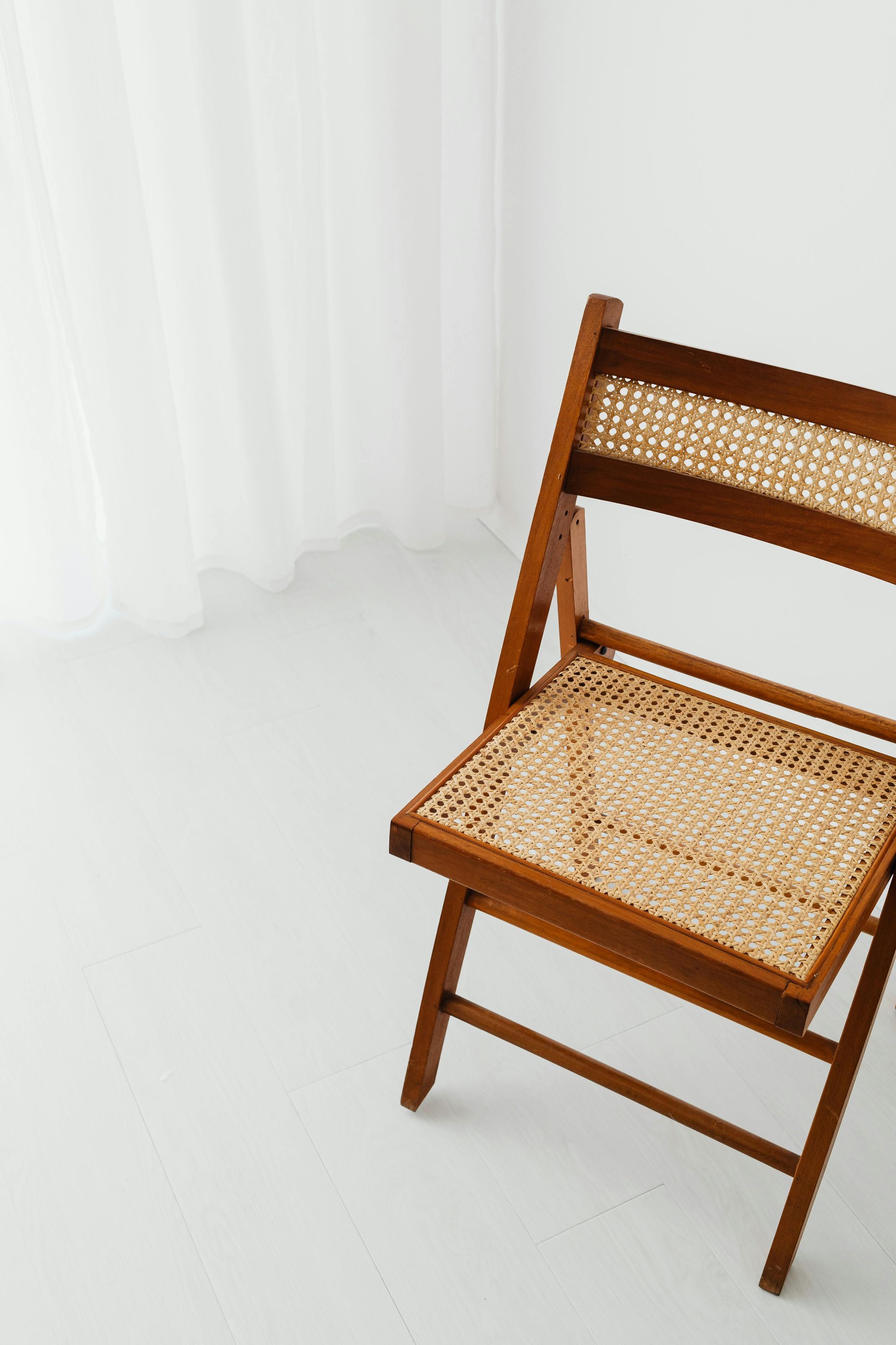 A wooden folding chair with a wicker seat is sitting on a white floor in front of a window.