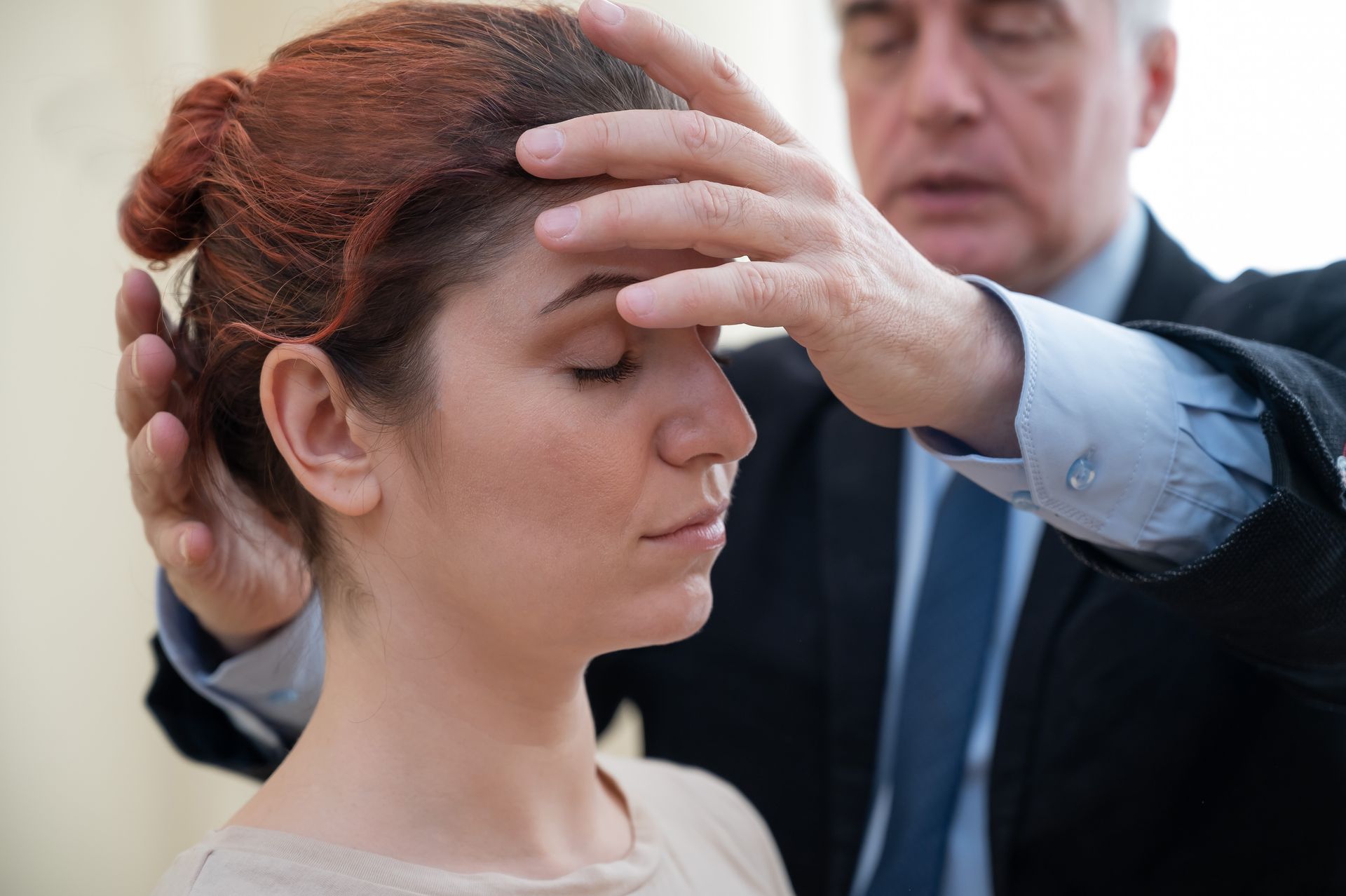 A woman with closed eyes stands calmly as a man places his hands on her forehead and head