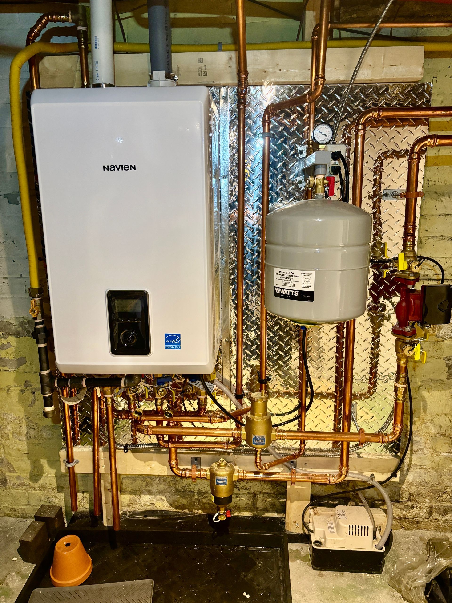 White wall-mounted boiler with copper pipes and expansion tank in a basement setting.