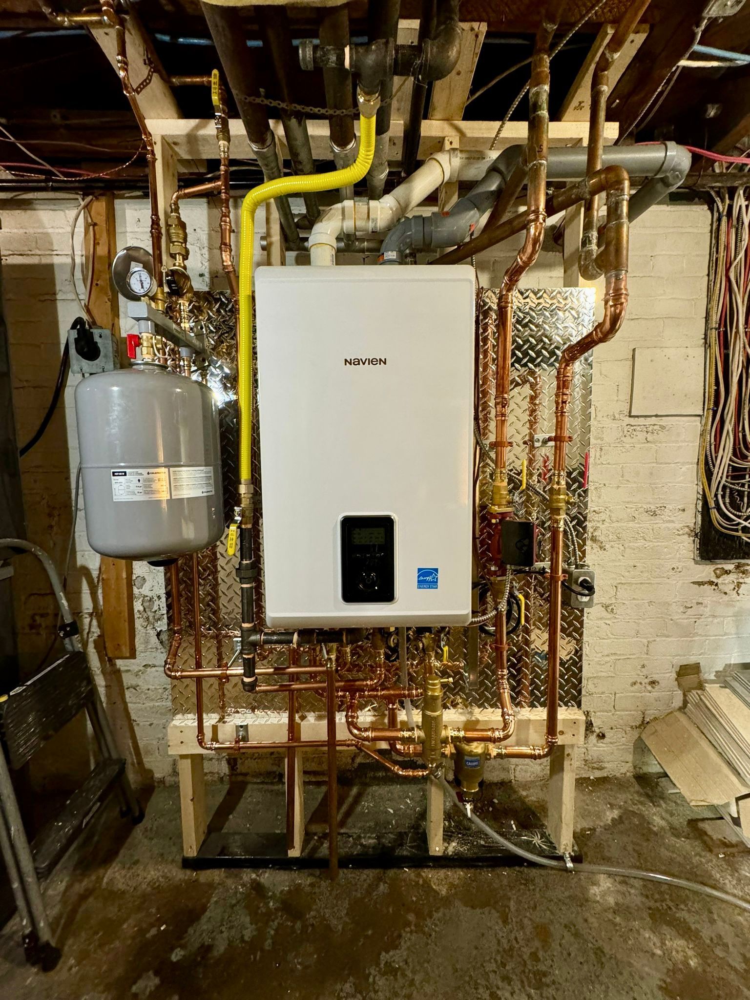Wall-mounted white boiler with copper pipes and a gray expansion tank in a basement setting.
