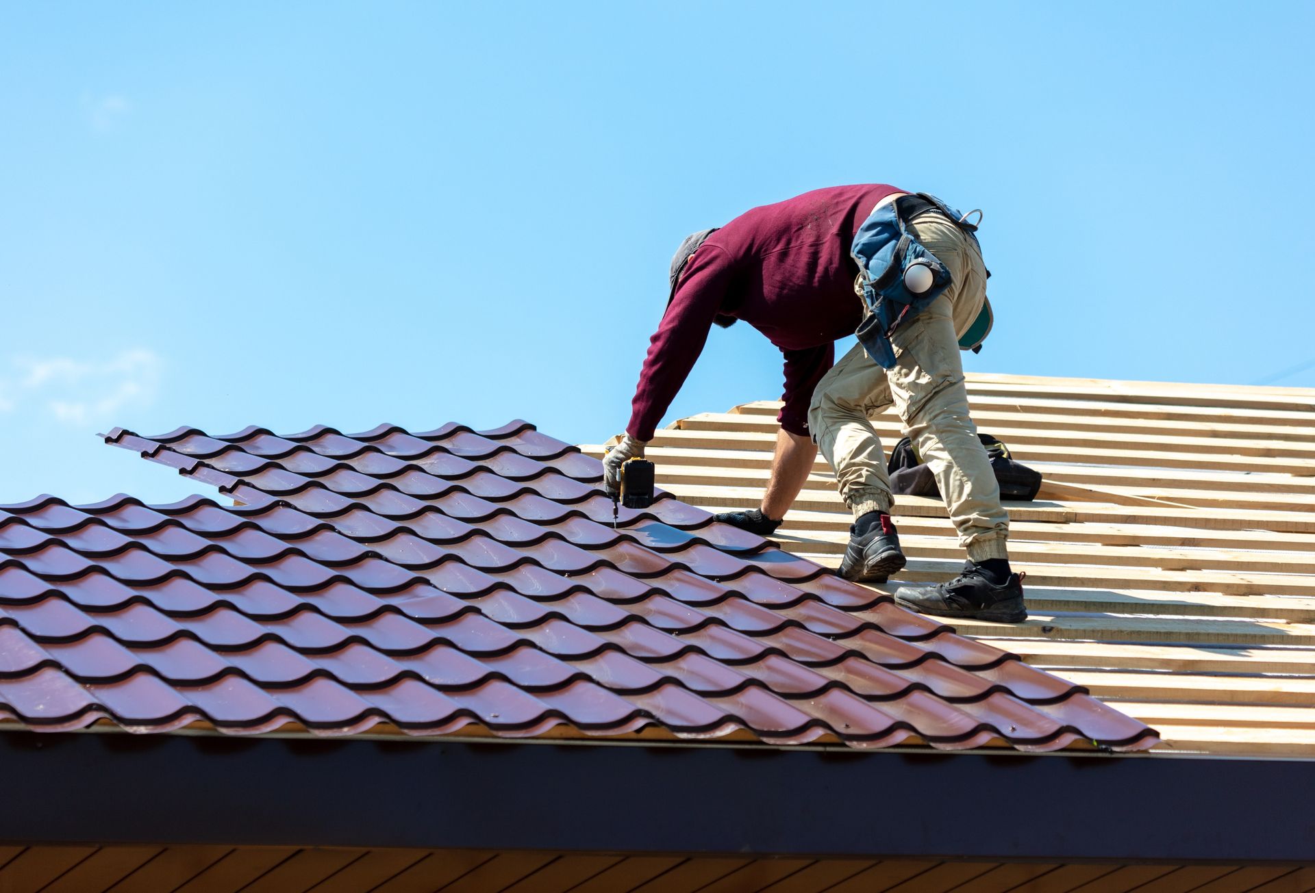 Worker roofing a house, installing red shingles beside a section of unfinished roof under a blue sky