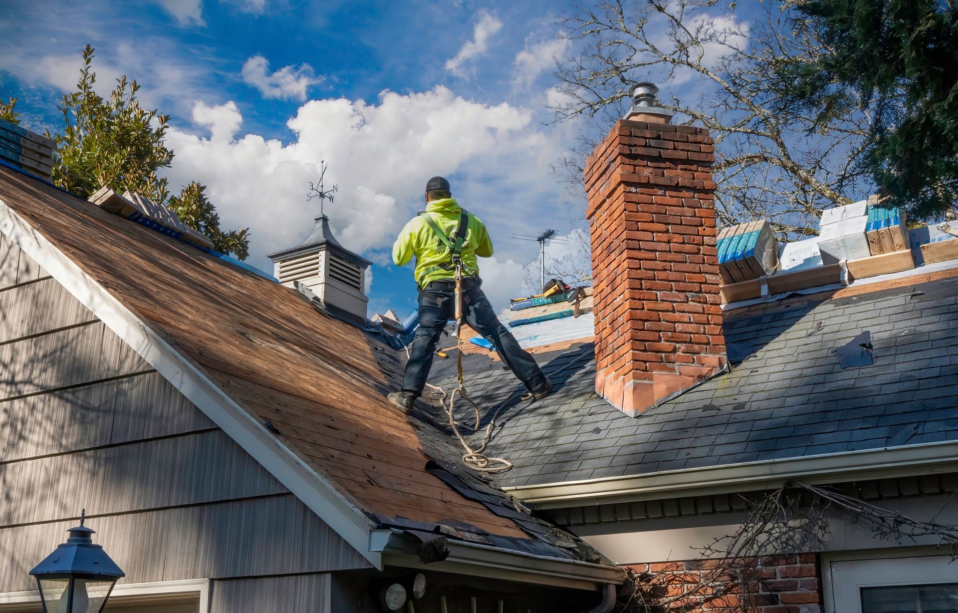 Roofing worker in a neon jacket on a sloped house roof near a brick chimney under a blue sky.
