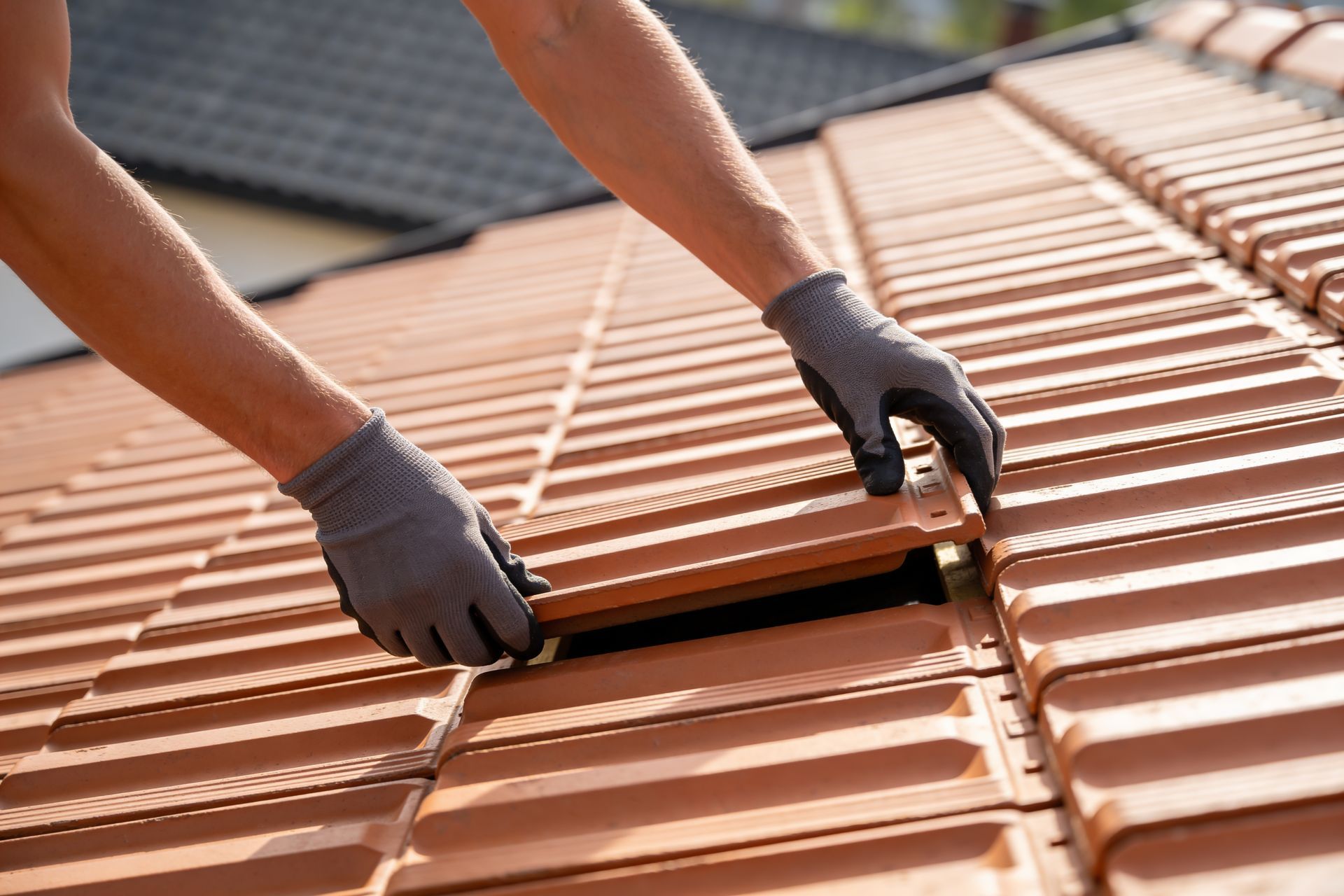 Gloved hands installing terracotta roof tiles on a roof with a partially removed tile at the center