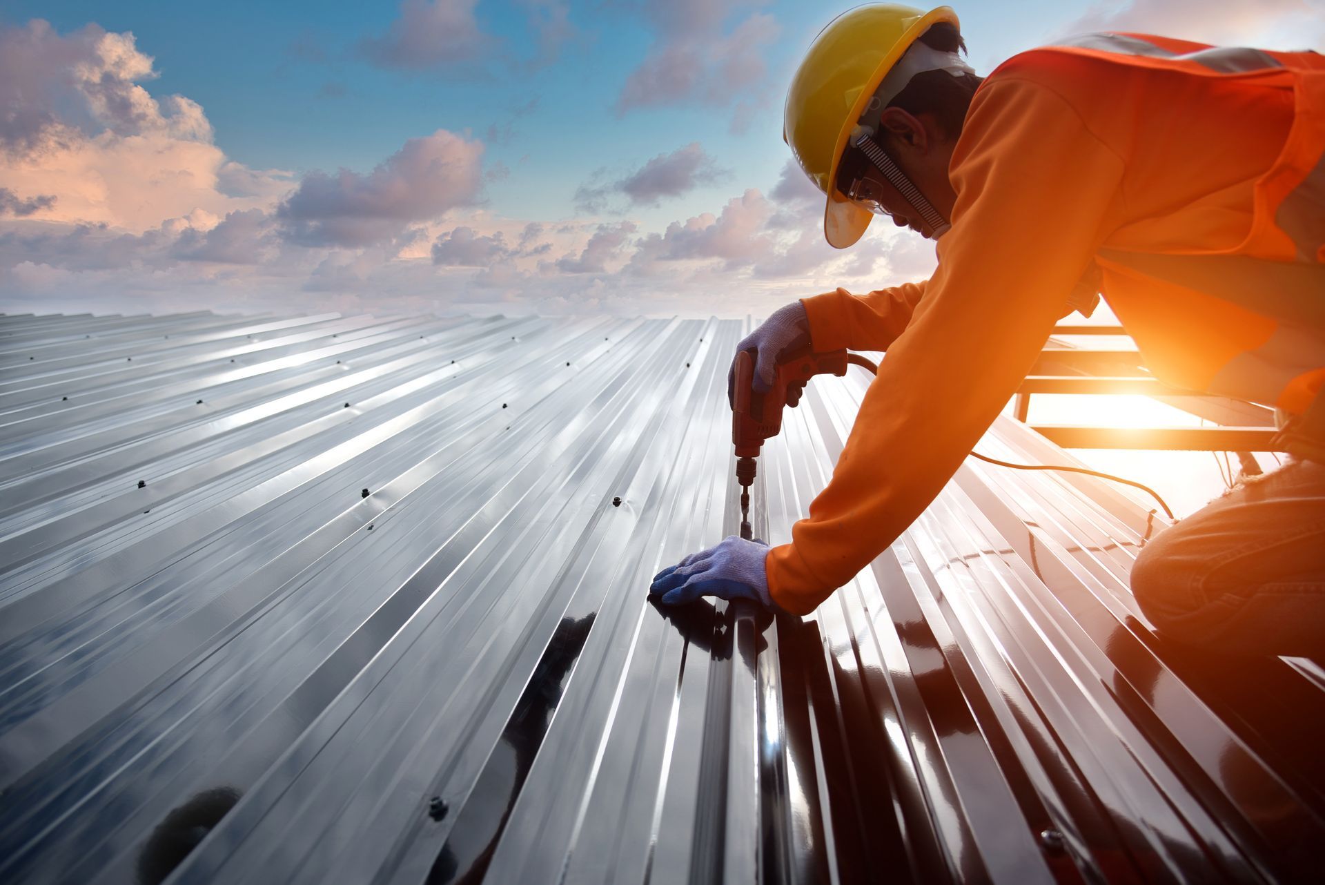 Worker in orange safety gear installing metal roofing panels at sunset