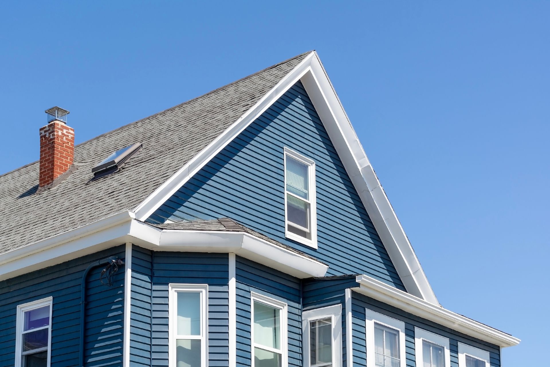 A blue house with white trim, a gable roof, a brick chimney, and a small skylight against a clear blue sky.