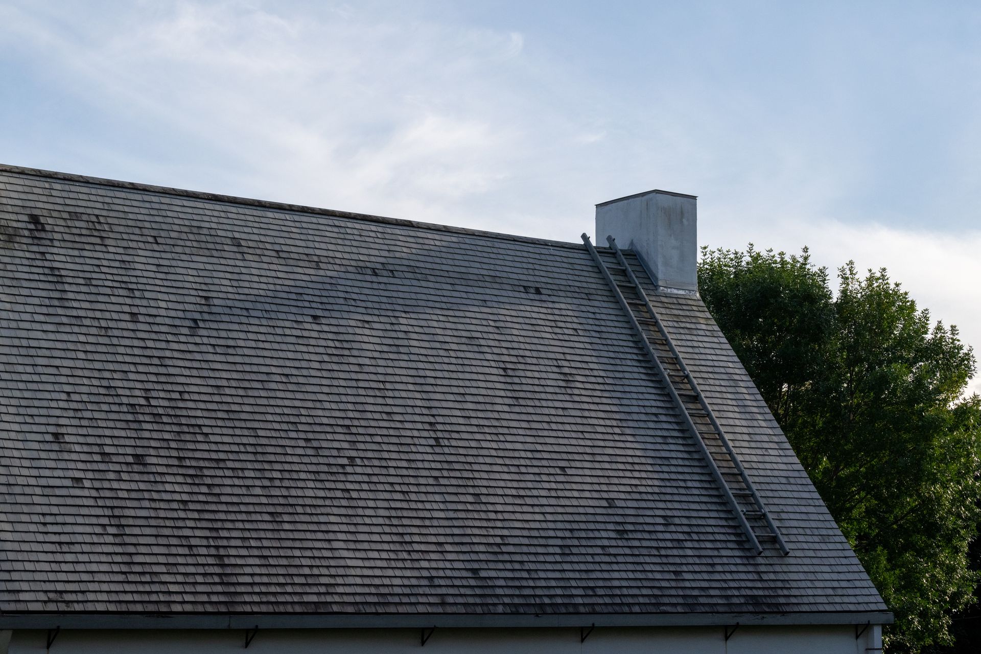 Gray shingled house roof with a chimney and tree against a cloudy sky