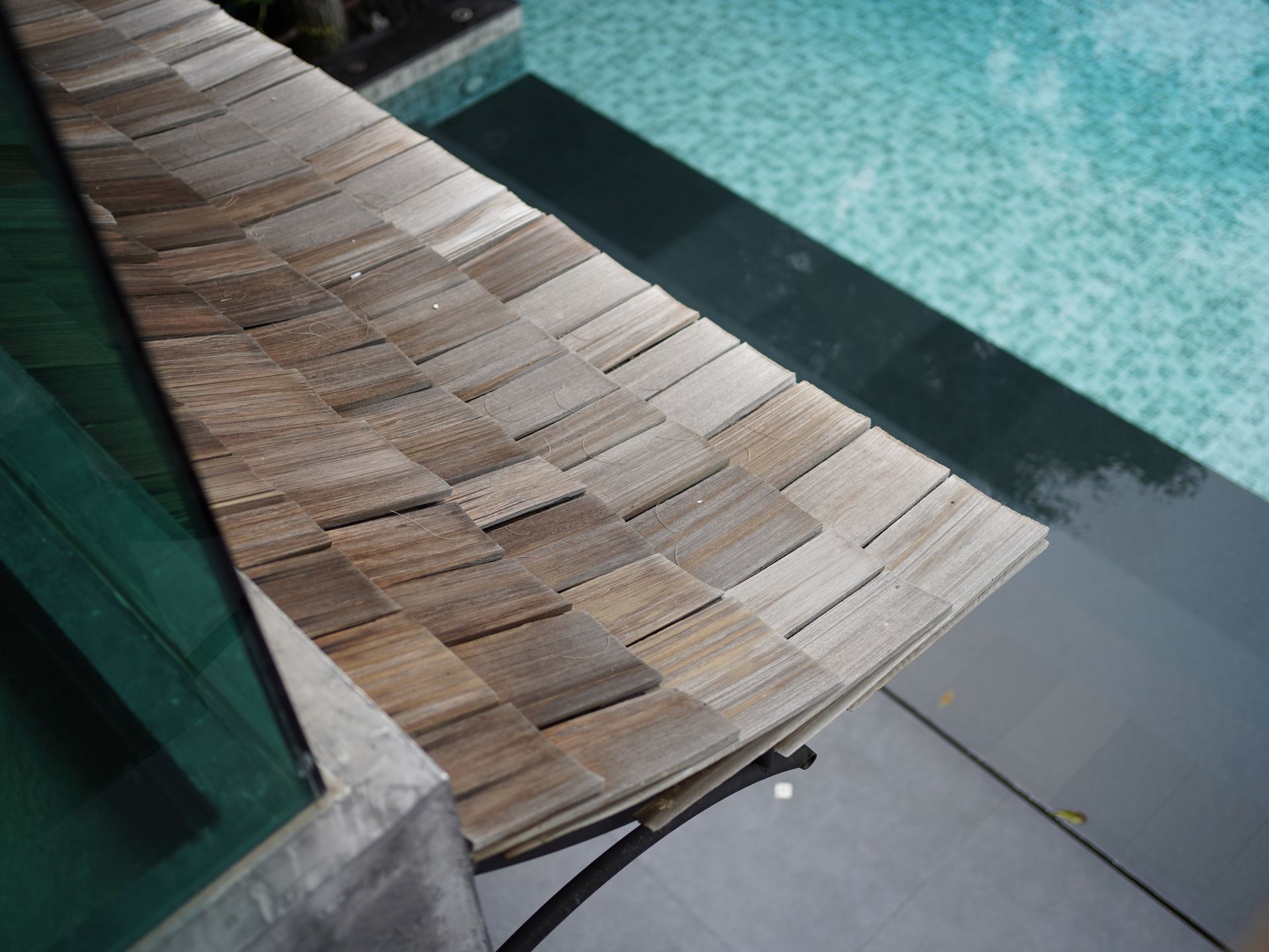 Woven pool lounge chair beside a turquoise swimming pool, with a dark walkway edge nearby