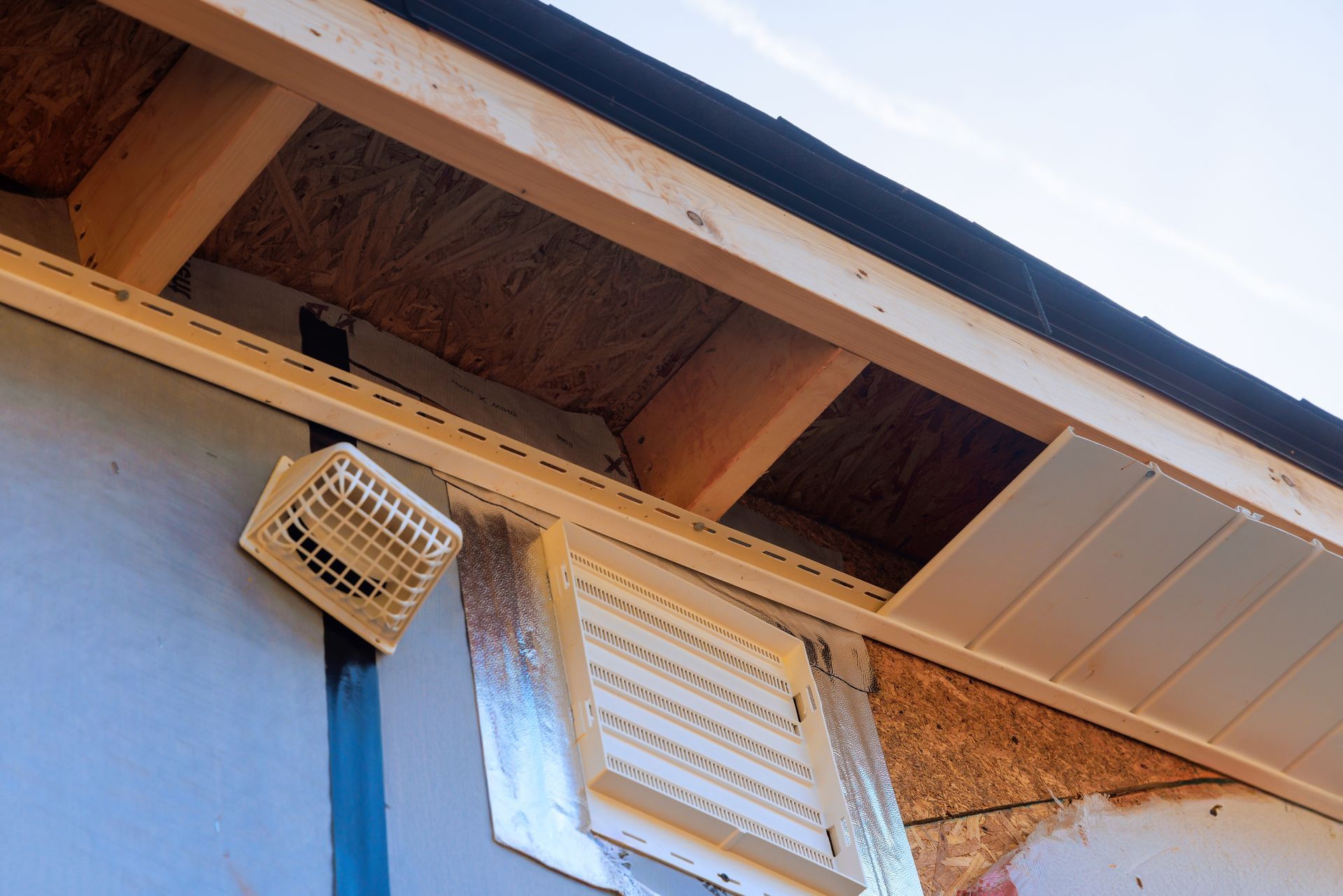 Corner of a house roof with exposed wooden eaves, a wall vent, and blue siding under bright sky