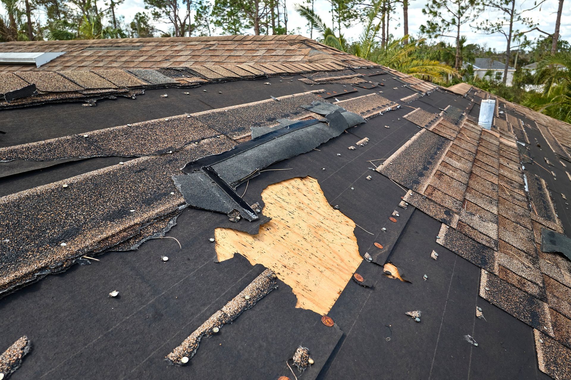 Damaged shingle roof with exposed plywood and scattered debris after storm damage