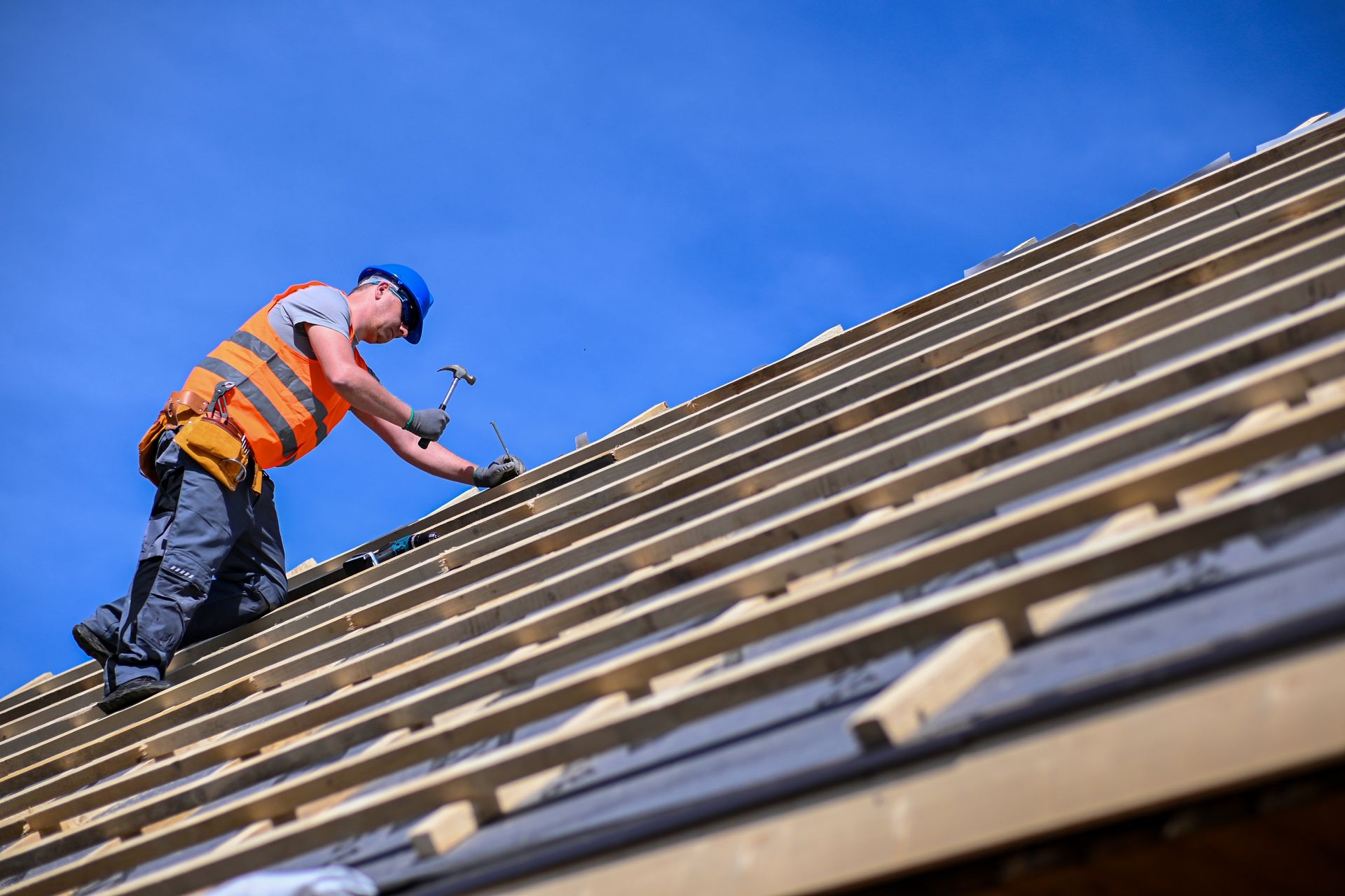 Construction worker in orange vest installing roof shingles on a steep rooftop under a blue sky