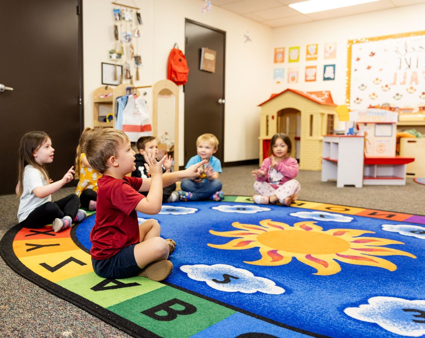 Children sitting in a circle on a rug, clapping, in a colorful classroom.