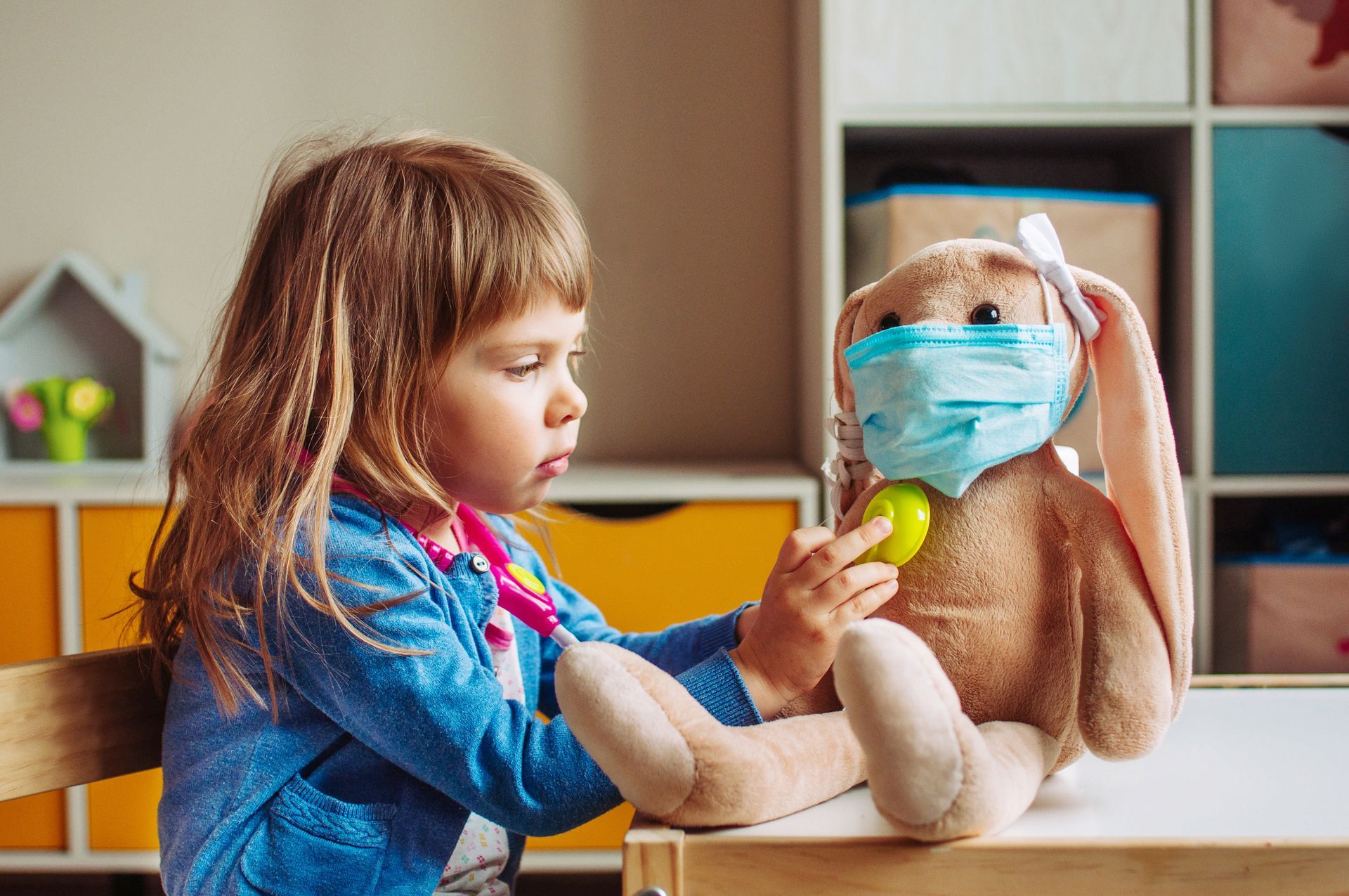 Young girl in blue shirt with stethoscope examines a plush bunny wearing a face mask at a table.