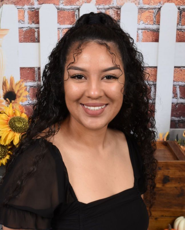 A woman with curly hair is smiling in front of a white picket fence.