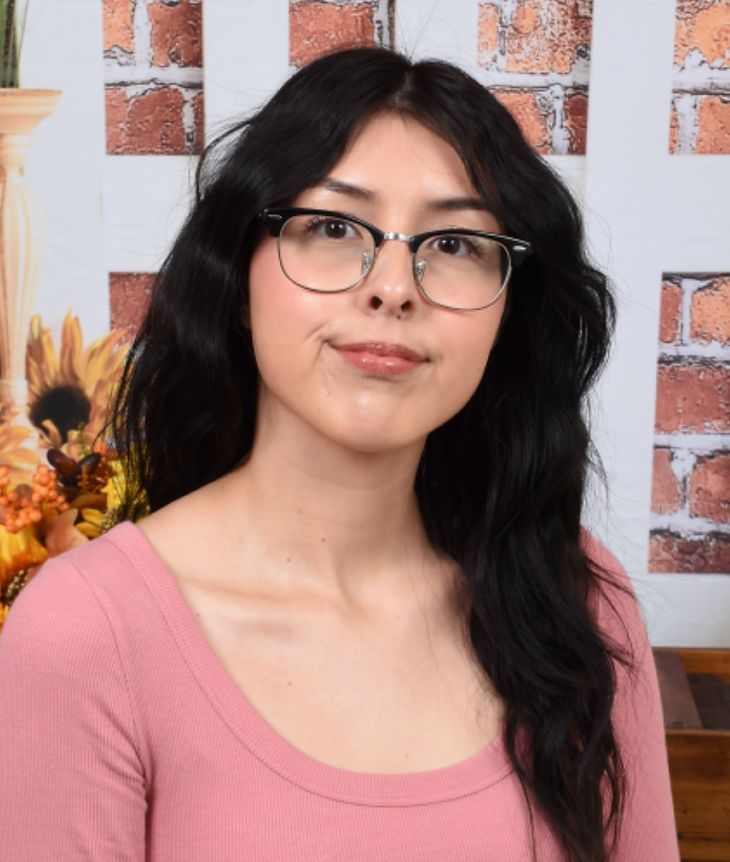 A woman wearing glasses and a pink shirt is standing in front of a brick wall.