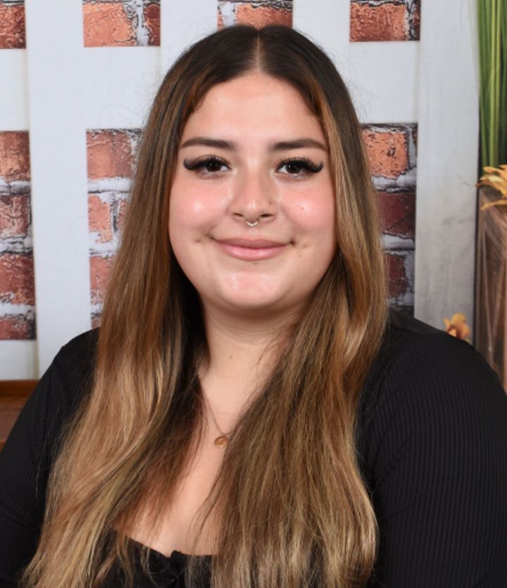 A woman with long hair and a nose ring is smiling in front of a brick wall.