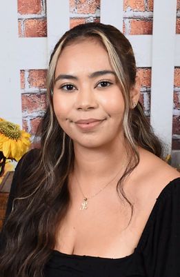 A woman wearing a black top and a necklace is standing in front of a brick wall.