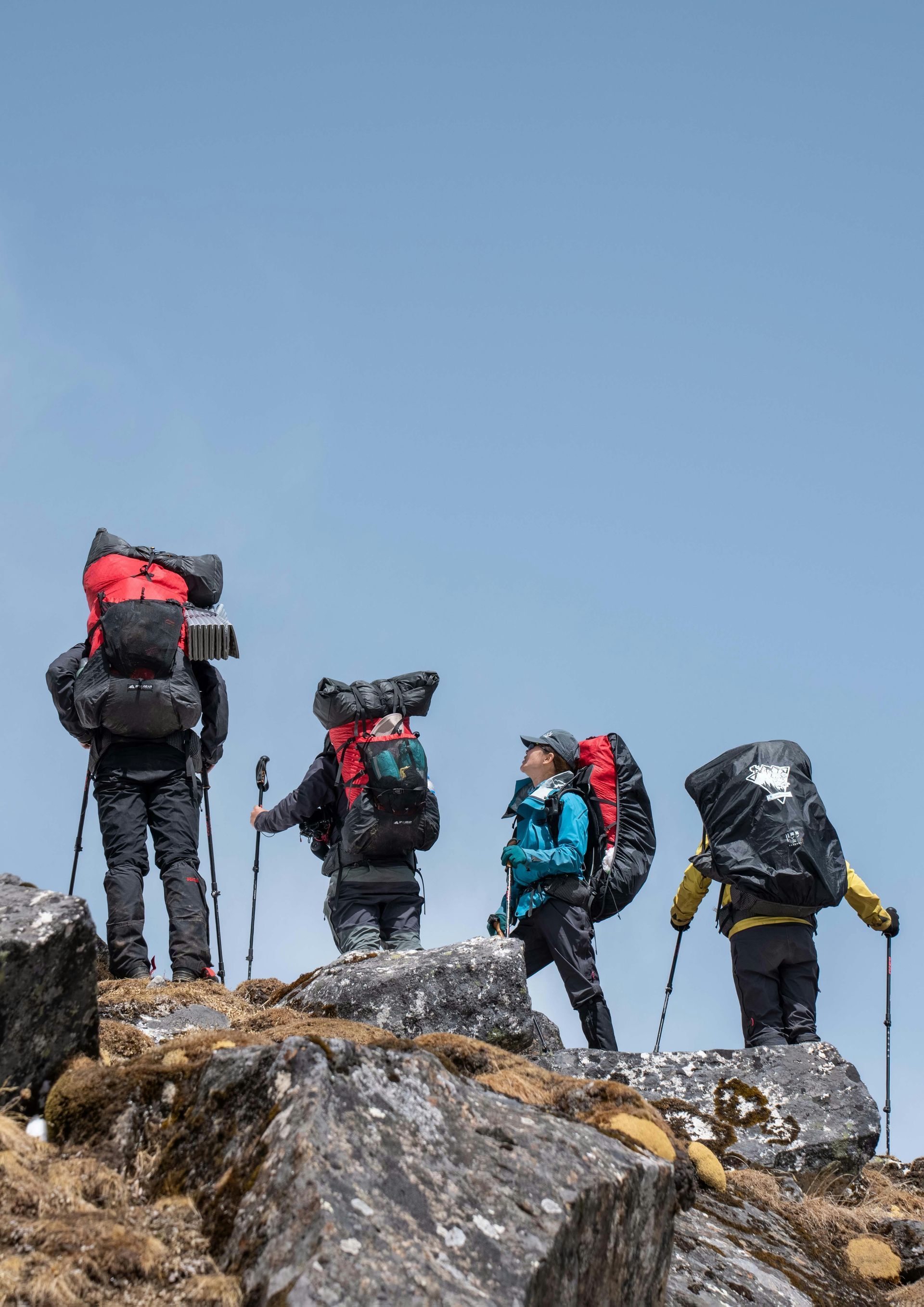 Quattro escursionisti con grossi zaini scalano un ripido pendio roccioso ad alta quota, sotto un cielo azzurro e limpido.