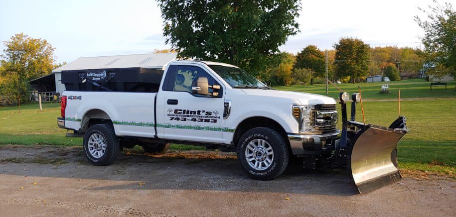 A white truck with a snow plow attached to it is parked in a parking lot.