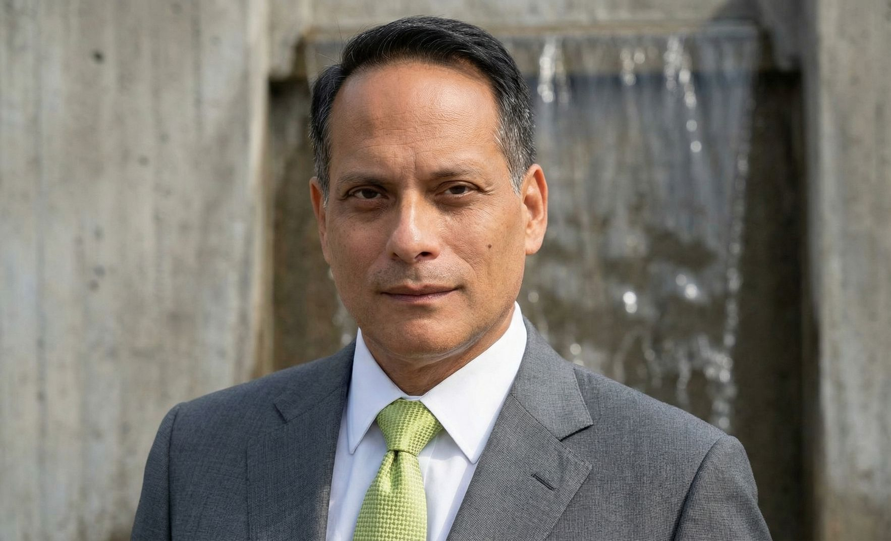 Headshot of a person in a grey suit and green tie, standing in front of a blurred outdoor waterfall.