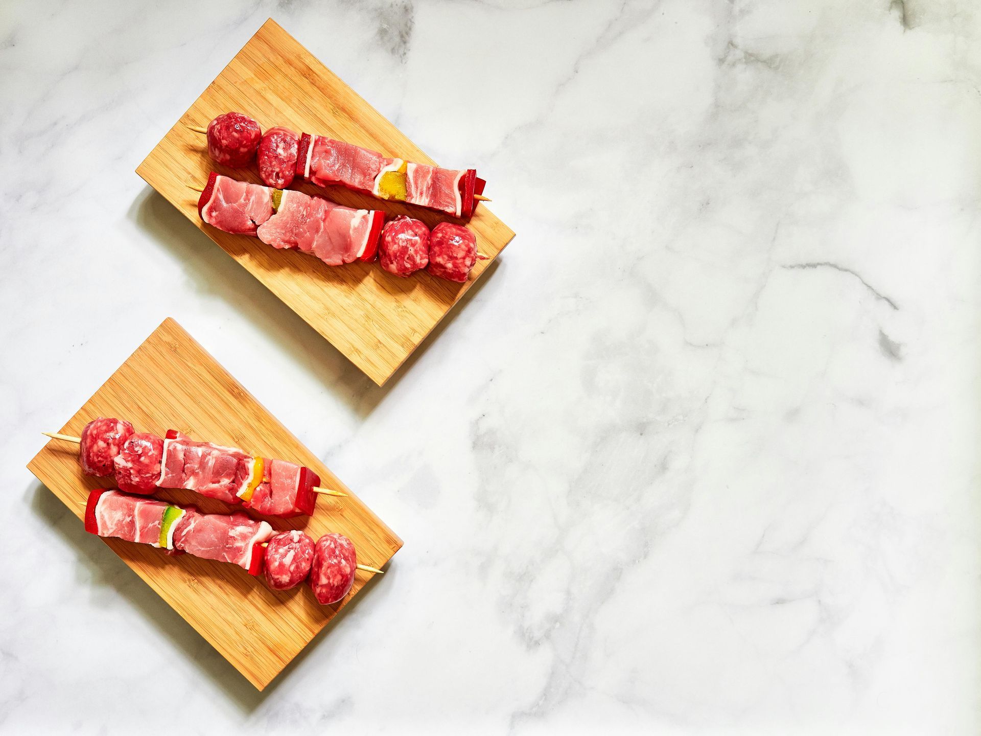 Two skewers of raw meat and vegetables on wooden boards, marble surface.