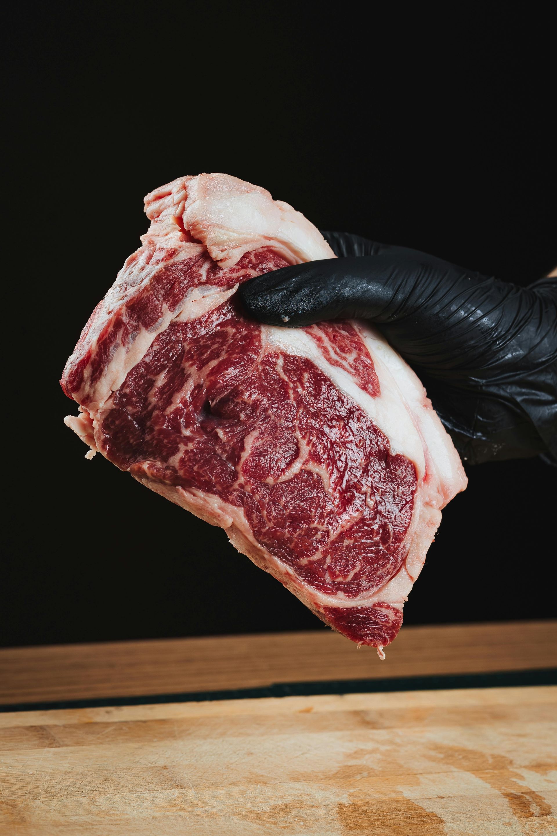 A gloved hand holds a thick, marbled raw steak against a black background, wooden surface.