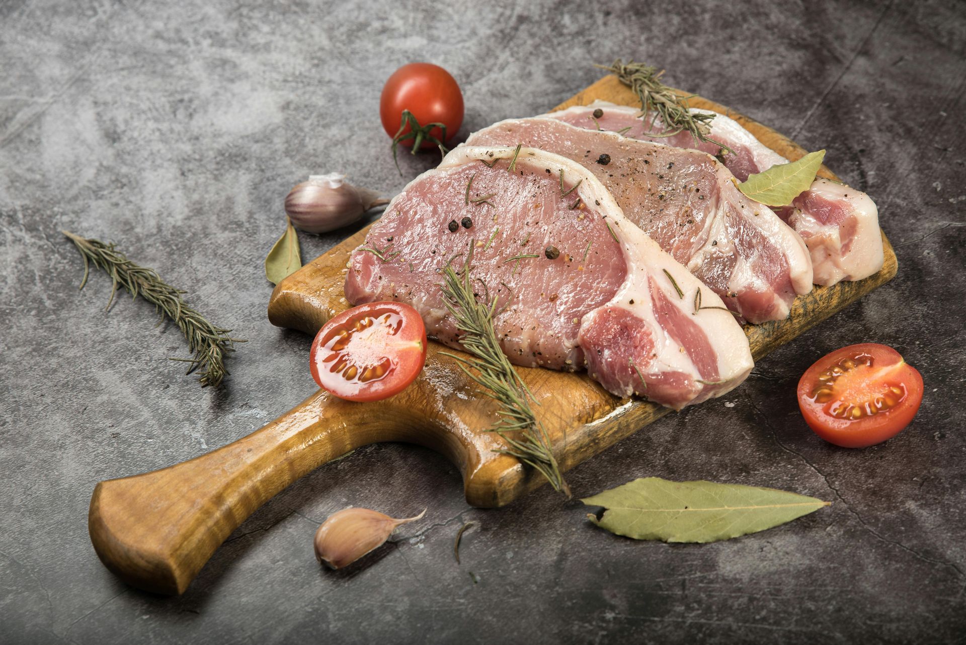Raw pork chops on a wooden board with herbs, tomatoes, and garlic, on a gray surface.
