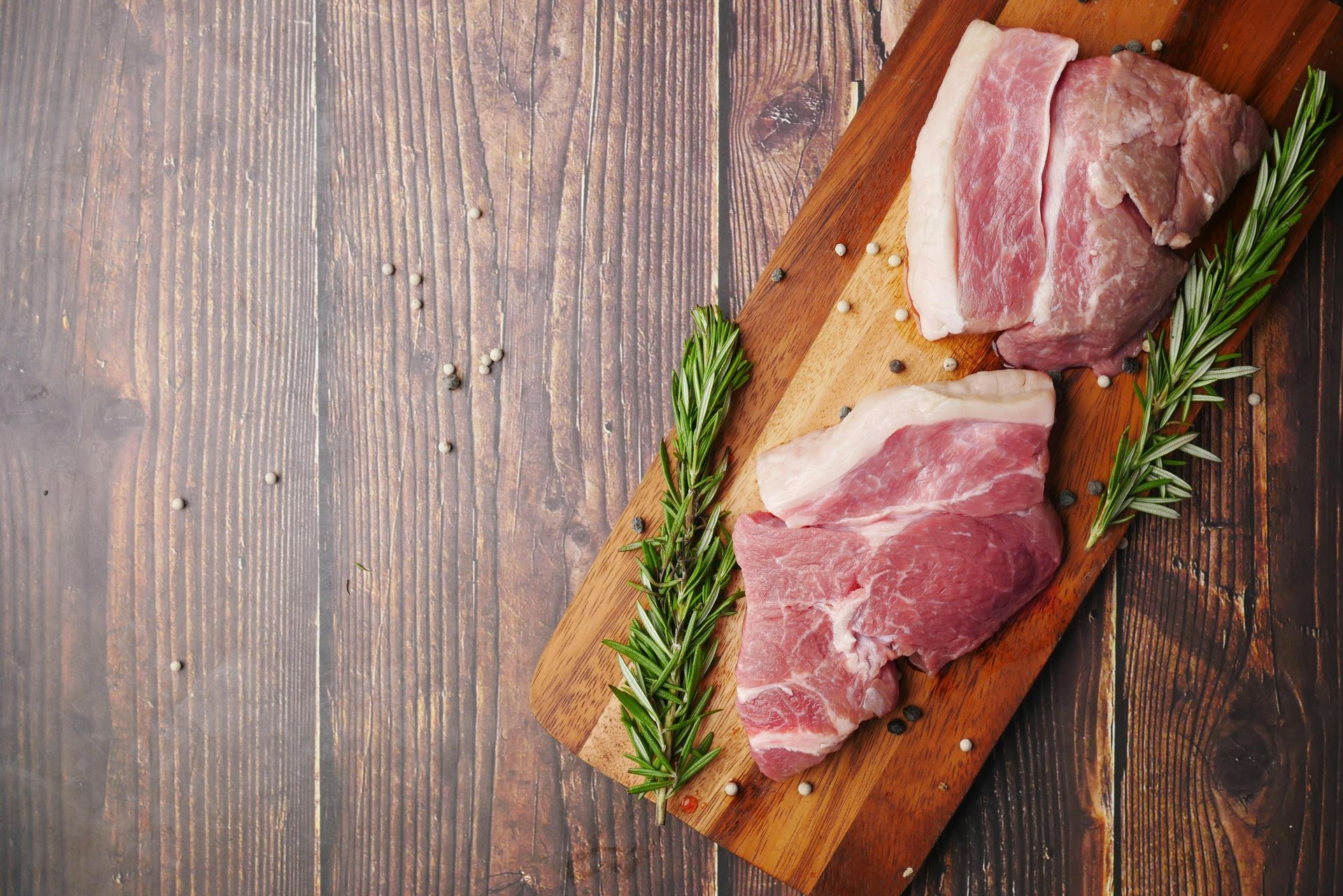 Raw meat on a wooden cutting board with rosemary sprigs, set on a wooden table.