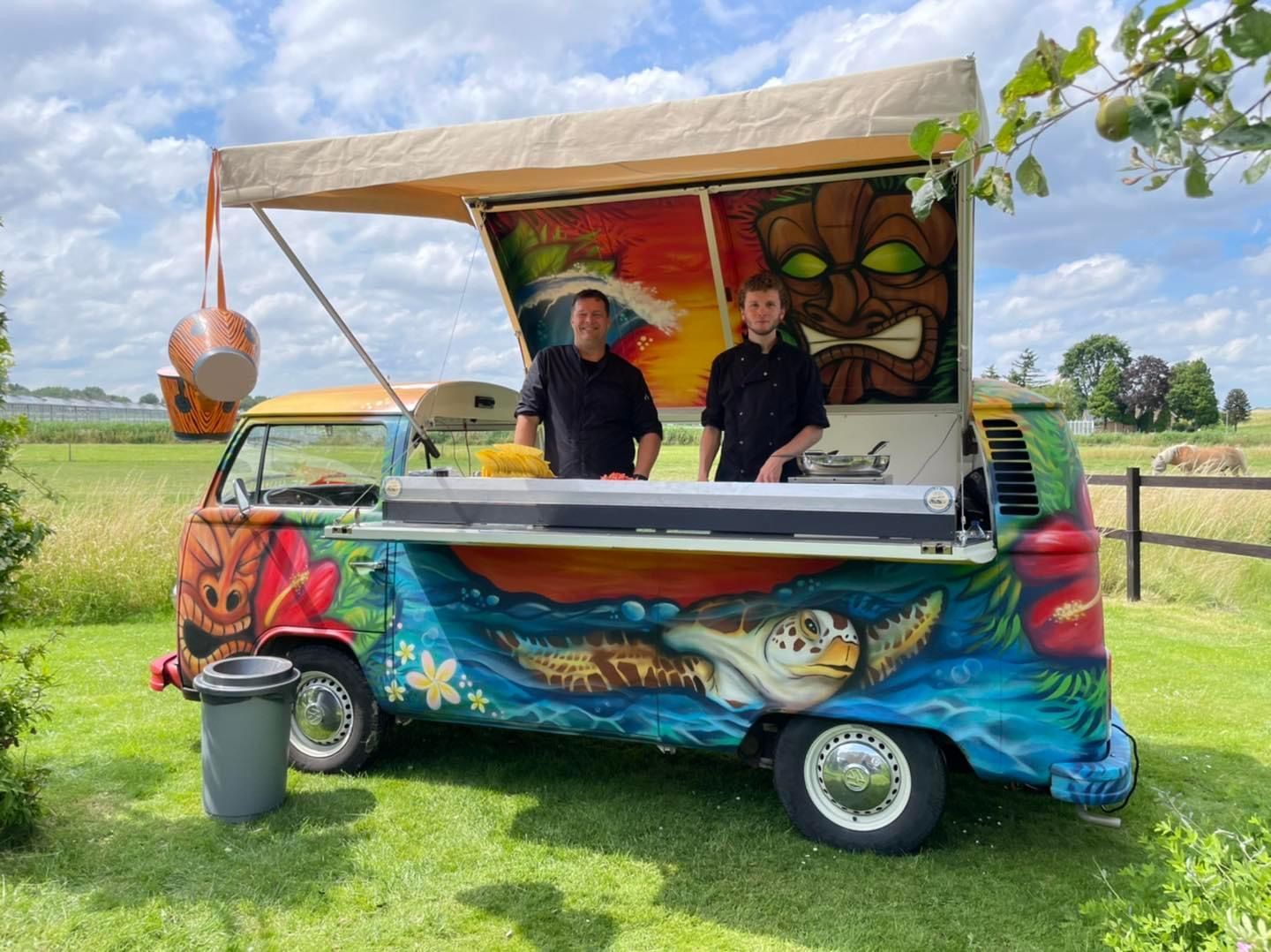 Twee mensen staan ​​achter een kleurrijke VW-bus met een eetkraam en tropische kunst.