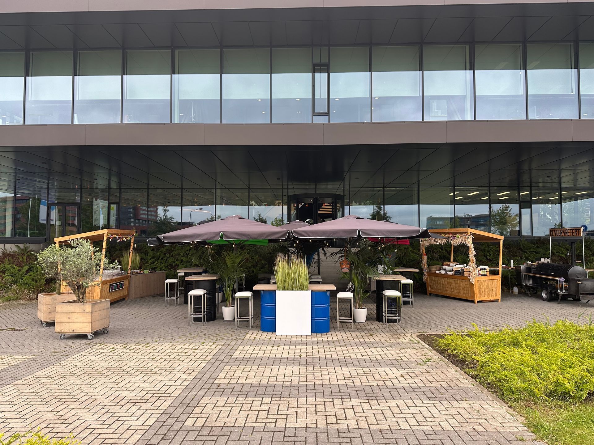 Een terras met tafels en parasols voor een modern gebouw. Er zijn foodtrucks en planten te zien.