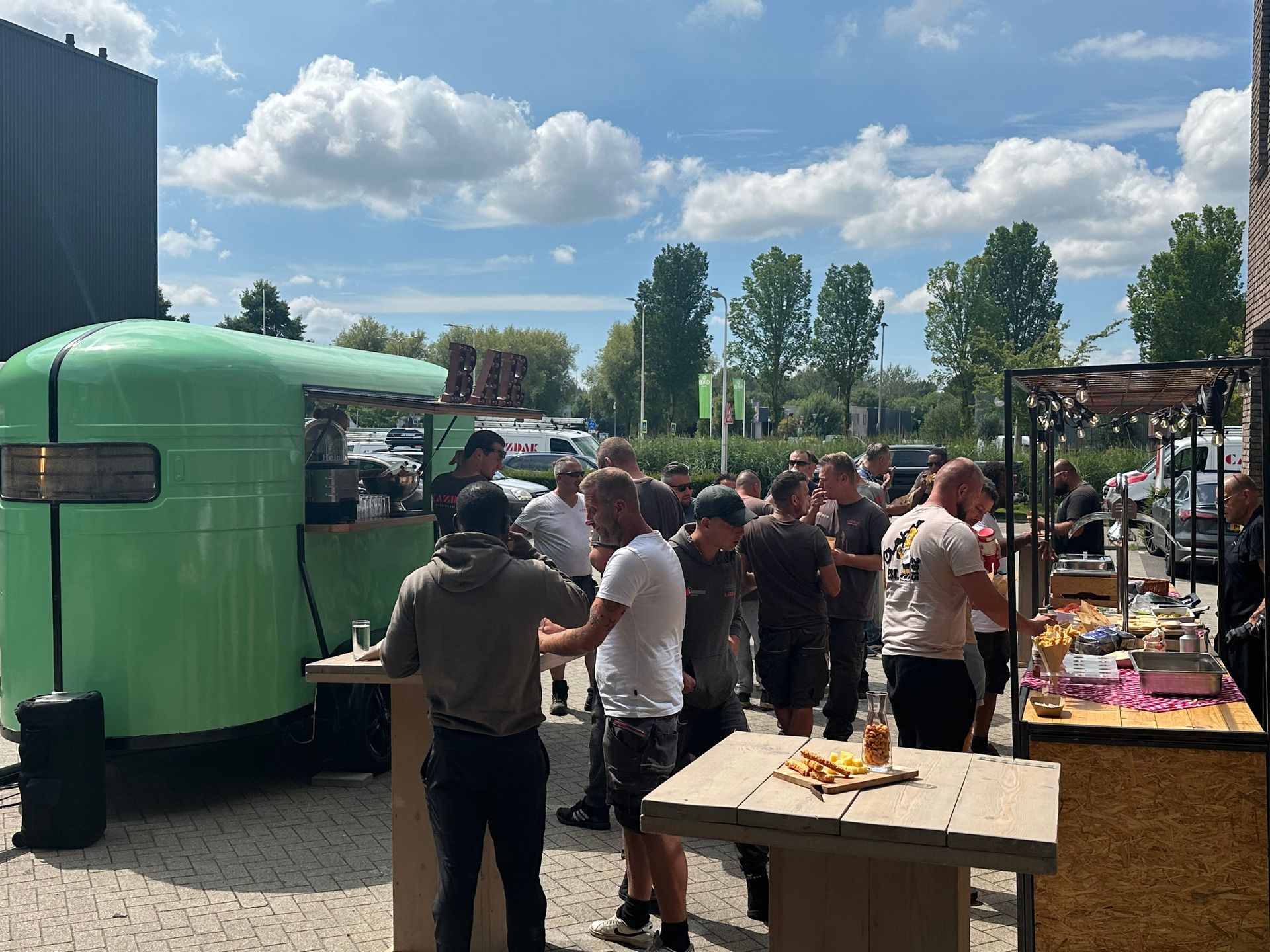 Op een zonnige dag verzamelen mensen zich buiten bij een groene foodtruck en een tafel met eten en drinken.